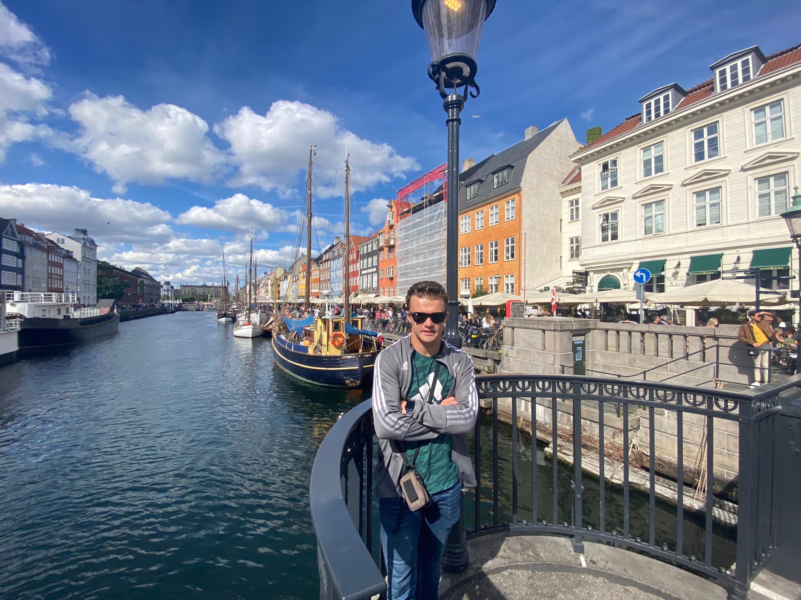 Nyhavn Copenhagen travel guide review, traveler standing on a bridge overlooking the colorful Nyhavn harbor with historic ships, waterfront cafés, and a busy crowd on a sunny day in Copenhagen.