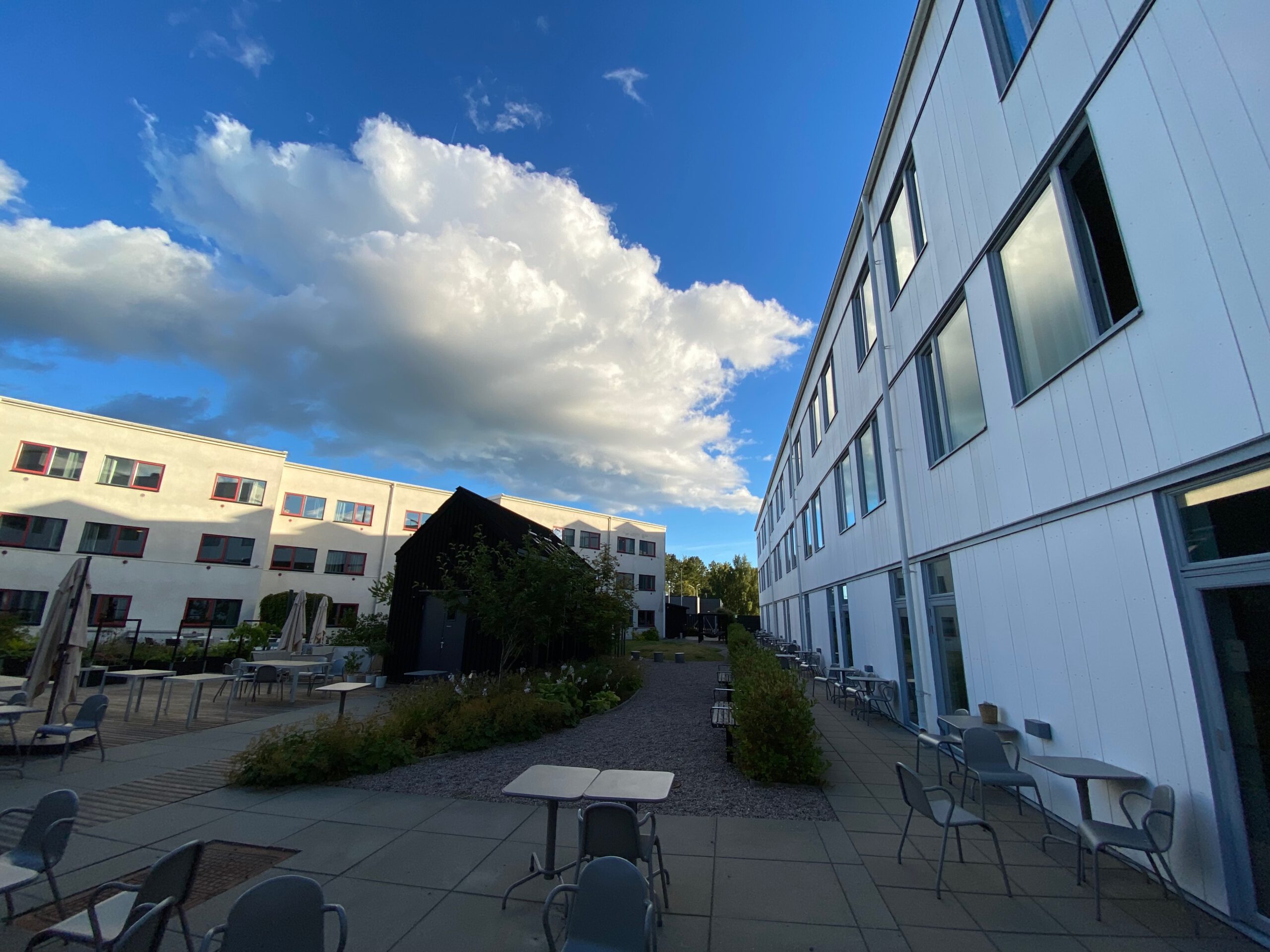 Exterior view of IKEA Hotell Älmhult showing multiple hotel buildings, outdoor seating areas, garden paths and a dramatic blue sky with large white clouds.