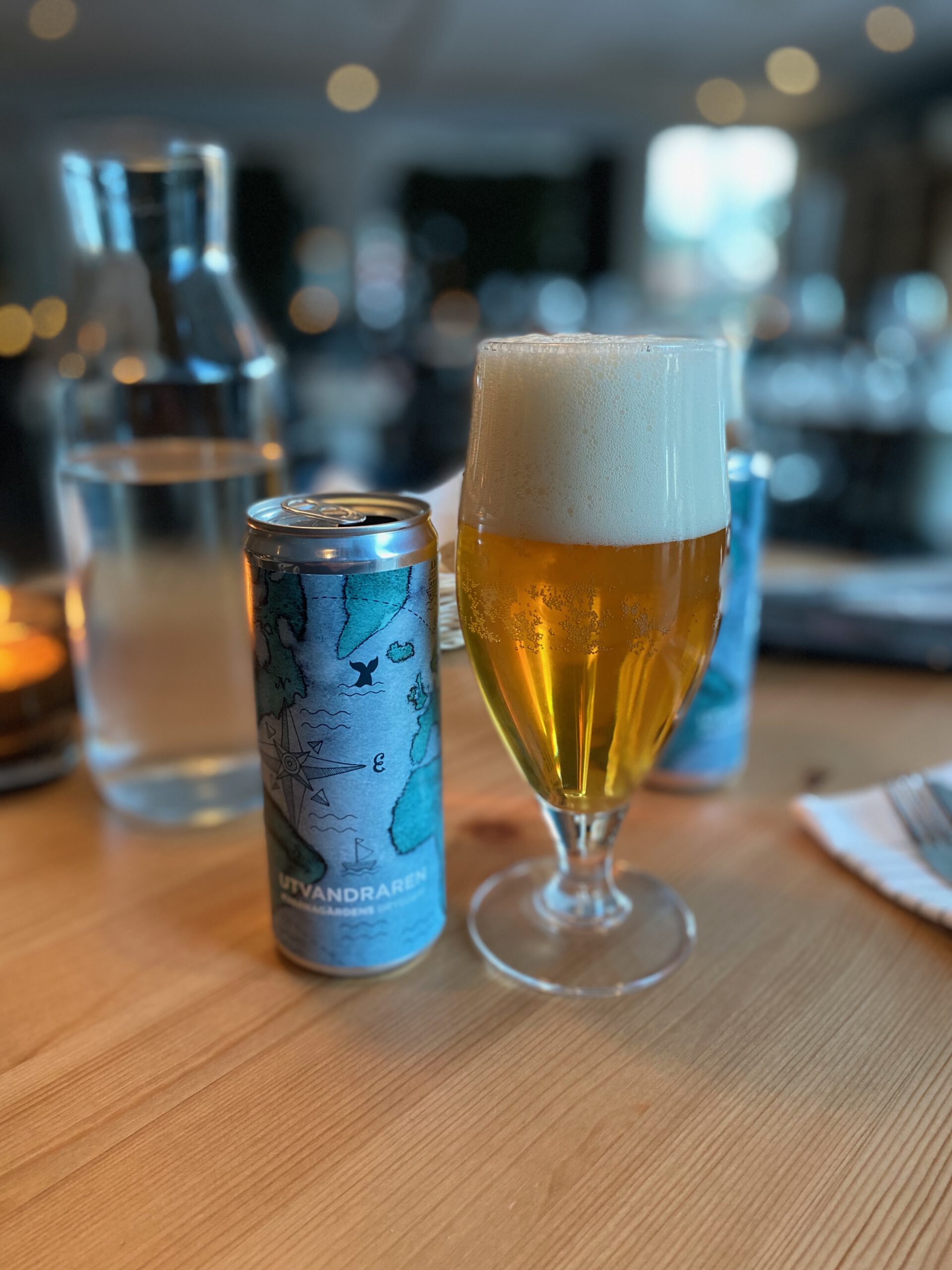 IKEA Hotell Restaurant Älmhult local Swedish craft beer Utvandaren, a clear golden lager style beer with a thick white foam head, served in a classic beer glass next to the original can on a wooden restaurant table, with soft indoor lighting and dining area in the background