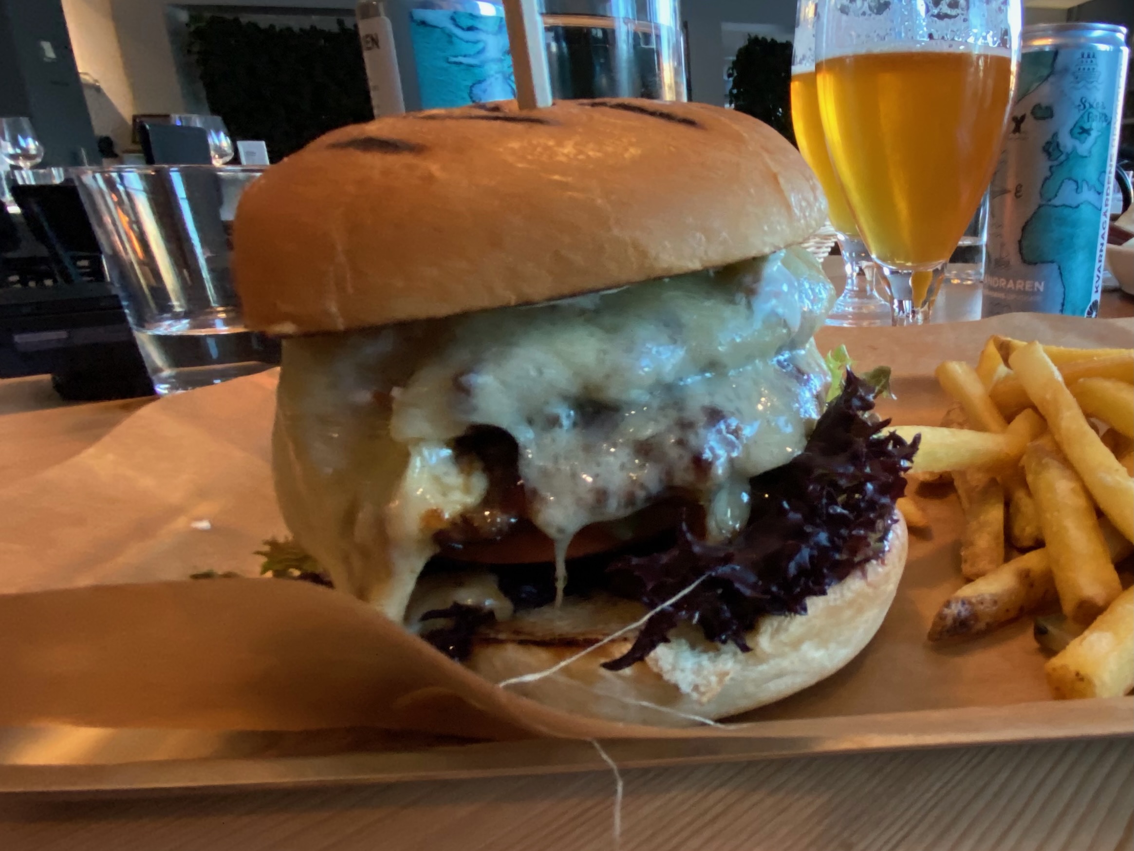 IKEA Hotell Restaurant Älmhult burger with grilled bun, juicy beef patty, melted cheese dripping over the sides, fresh lettuce and thick fries served on baking paper with a glass of local Swedish beer in the background
