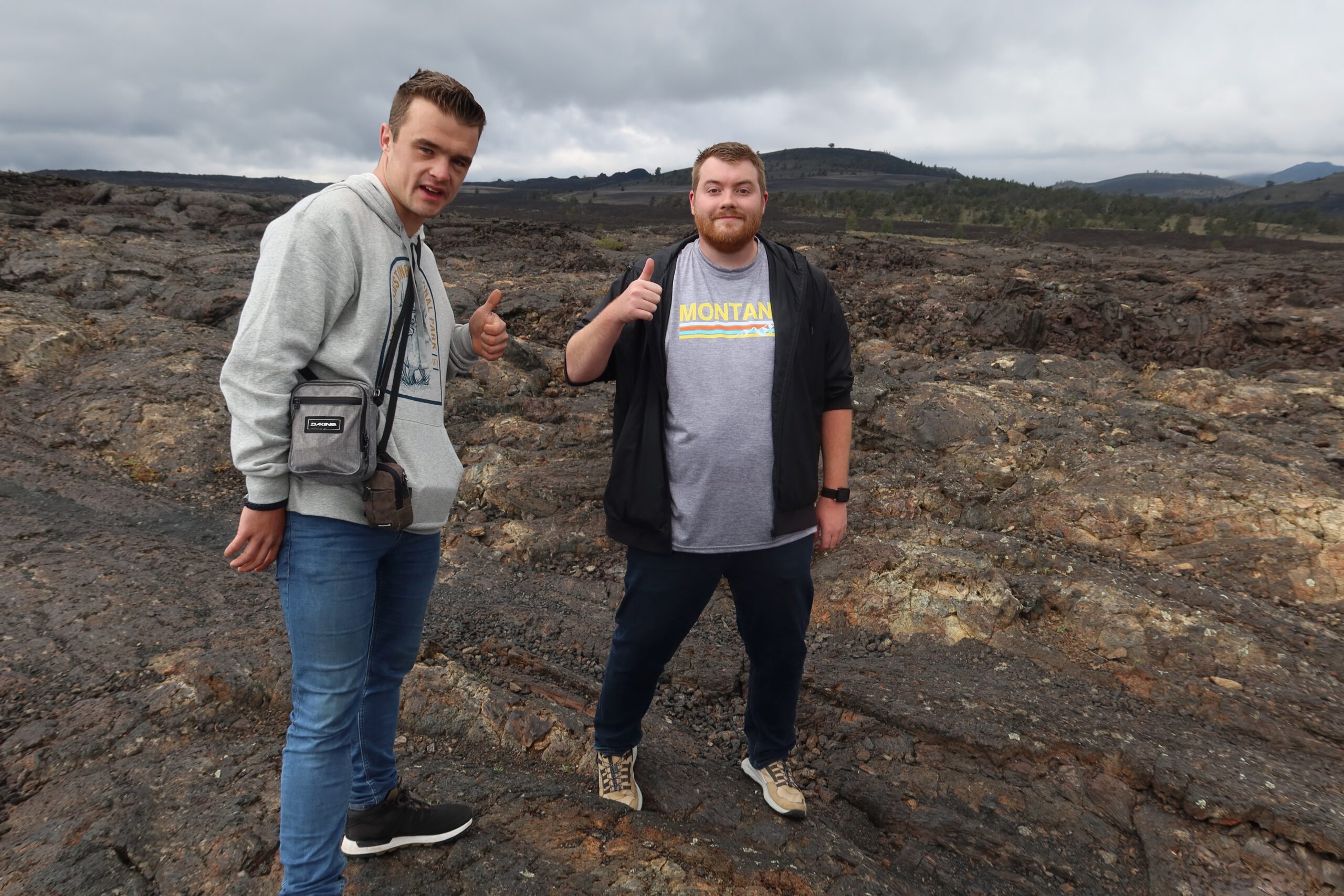 Two travelers standing on volcanic lava fields at Craters of the Moon National Monument in Idaho, giving thumbs up during a road trip stop
