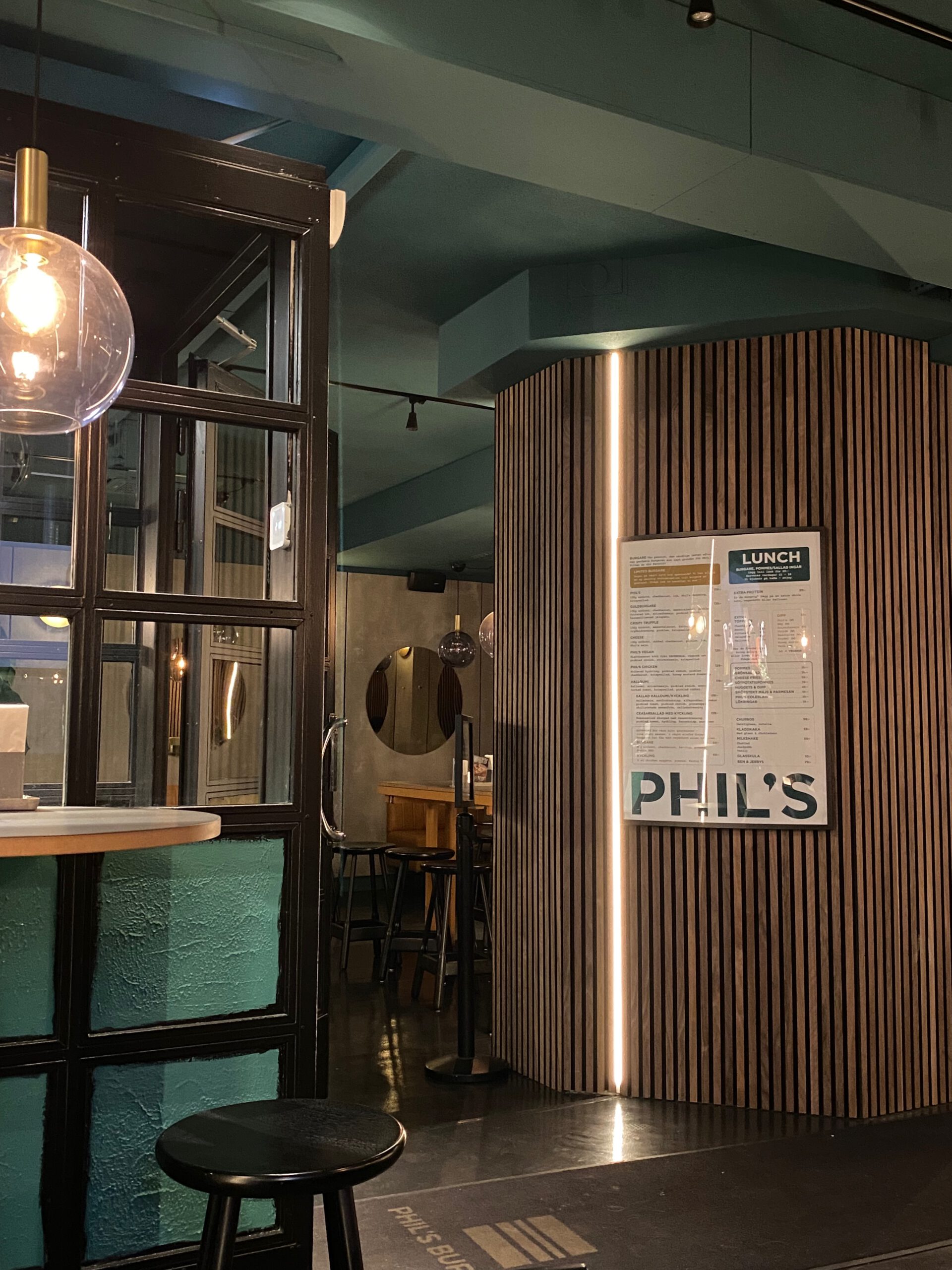 Interior of Phils Burger Drottninggatan Stockholm, showing the modern restaurant design with menu boards, green accents, wood paneling, and the dining area where burgers and Belgian-style beers are served