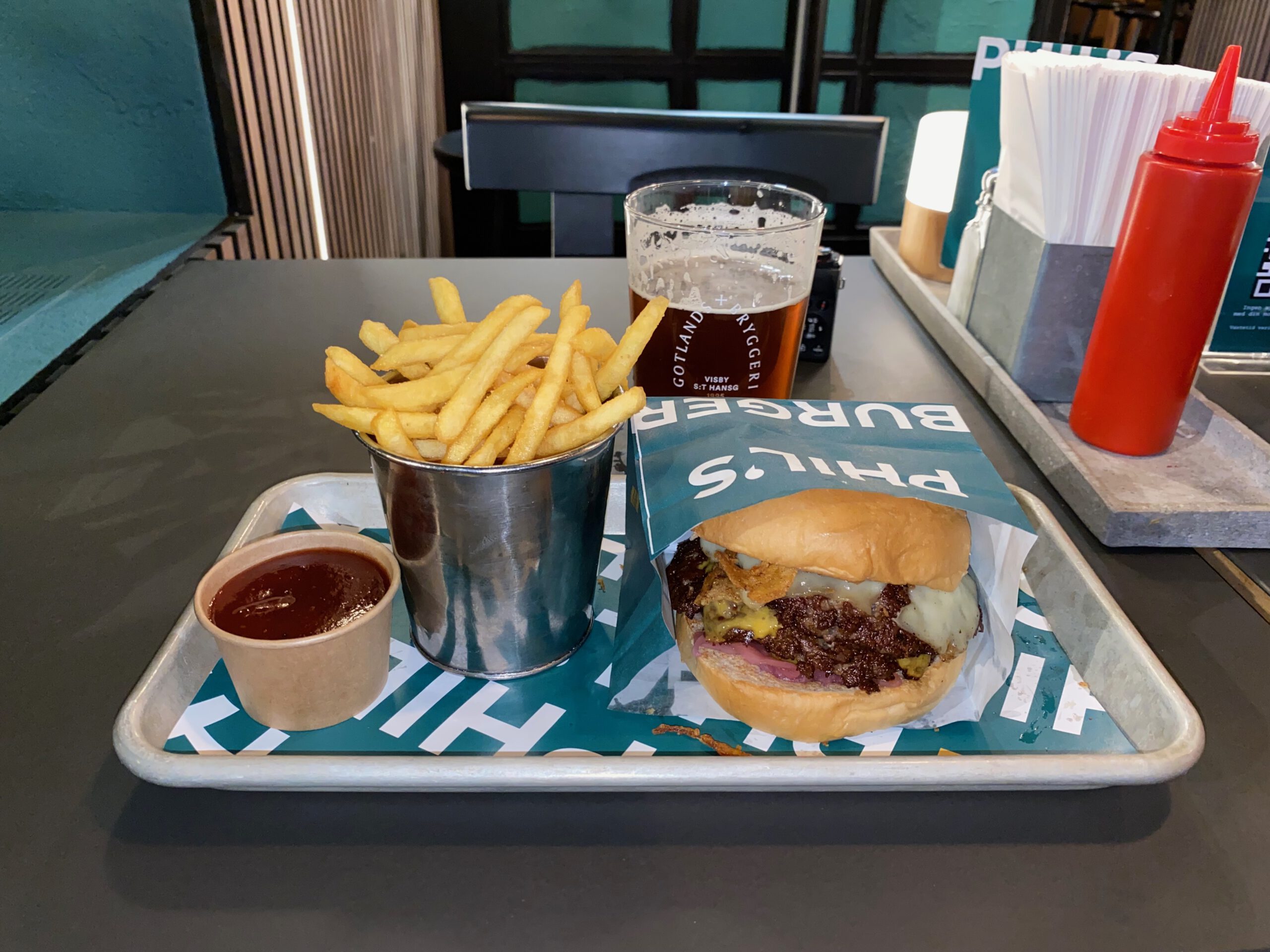 Phils Burger Drottninggatan Stockholm serving a juicy beef burger with melted cheese, caramelized onions and sauce in a soft bun, accompanied by crispy fries in a metal cup, barbecue-style dipping sauce, and a Swedish Gotlands Bryggeri craft beer on a tray inside the restaurant