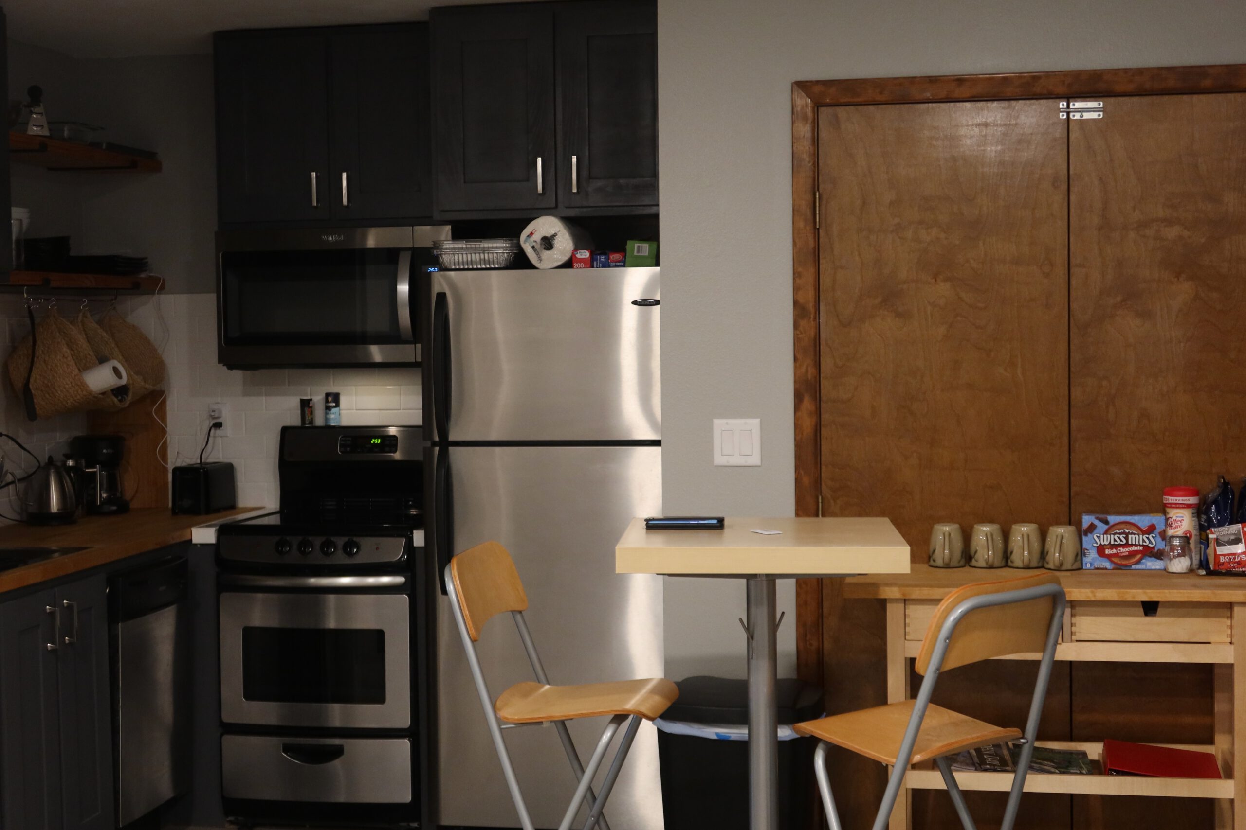 Compact kitchen inside the John Day Airbnb featuring dark cabinets, a stainless steel refrigerator and oven, open wooden shelves, and a small dining table with two chairs in a simple, modern layout.