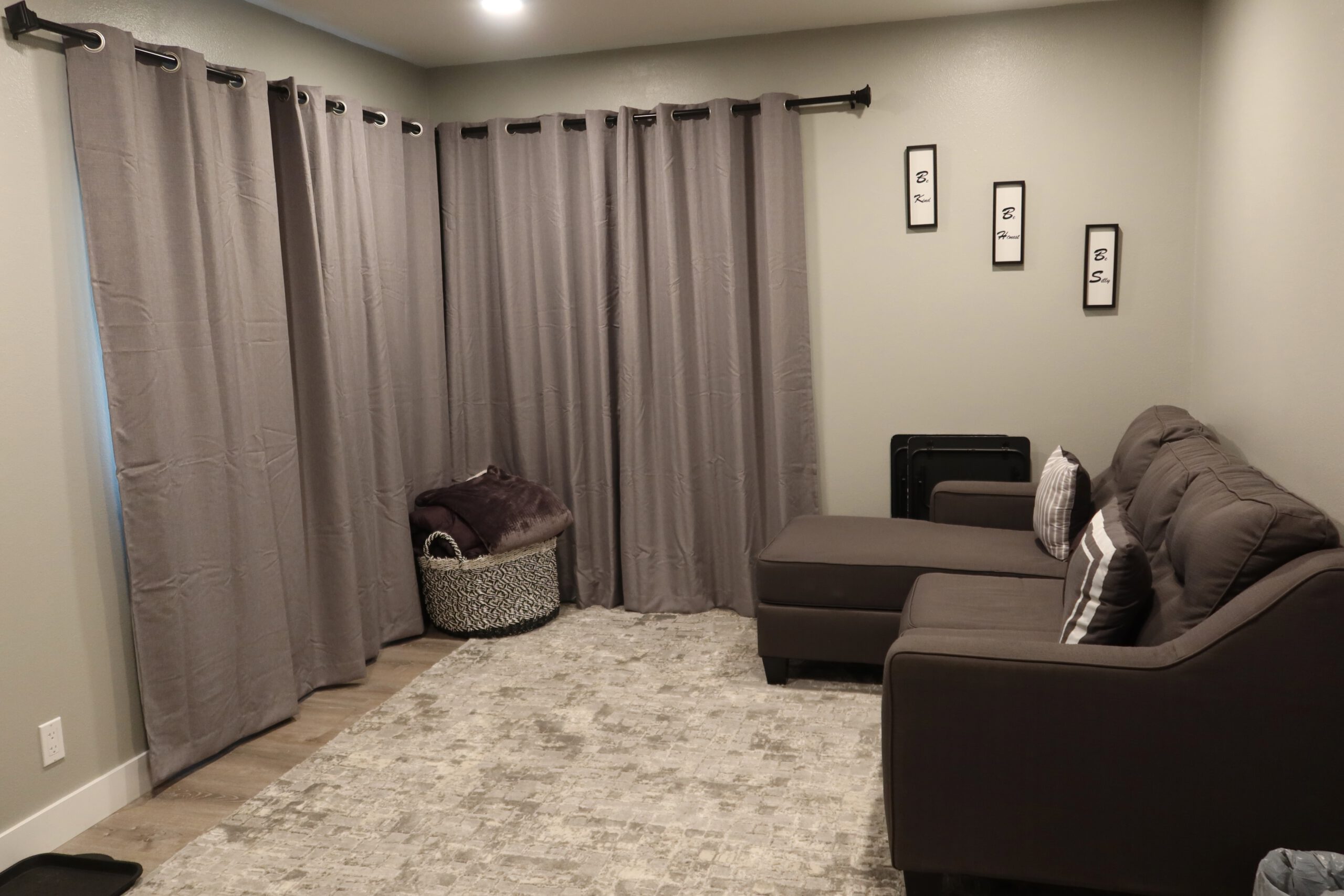 Living room inside the Airbnb John Day Oregon with a brown sectional sofa, blackout curtains covering the windows, soft area rug, and a basket with extra blankets creating a simple and functional seating area.