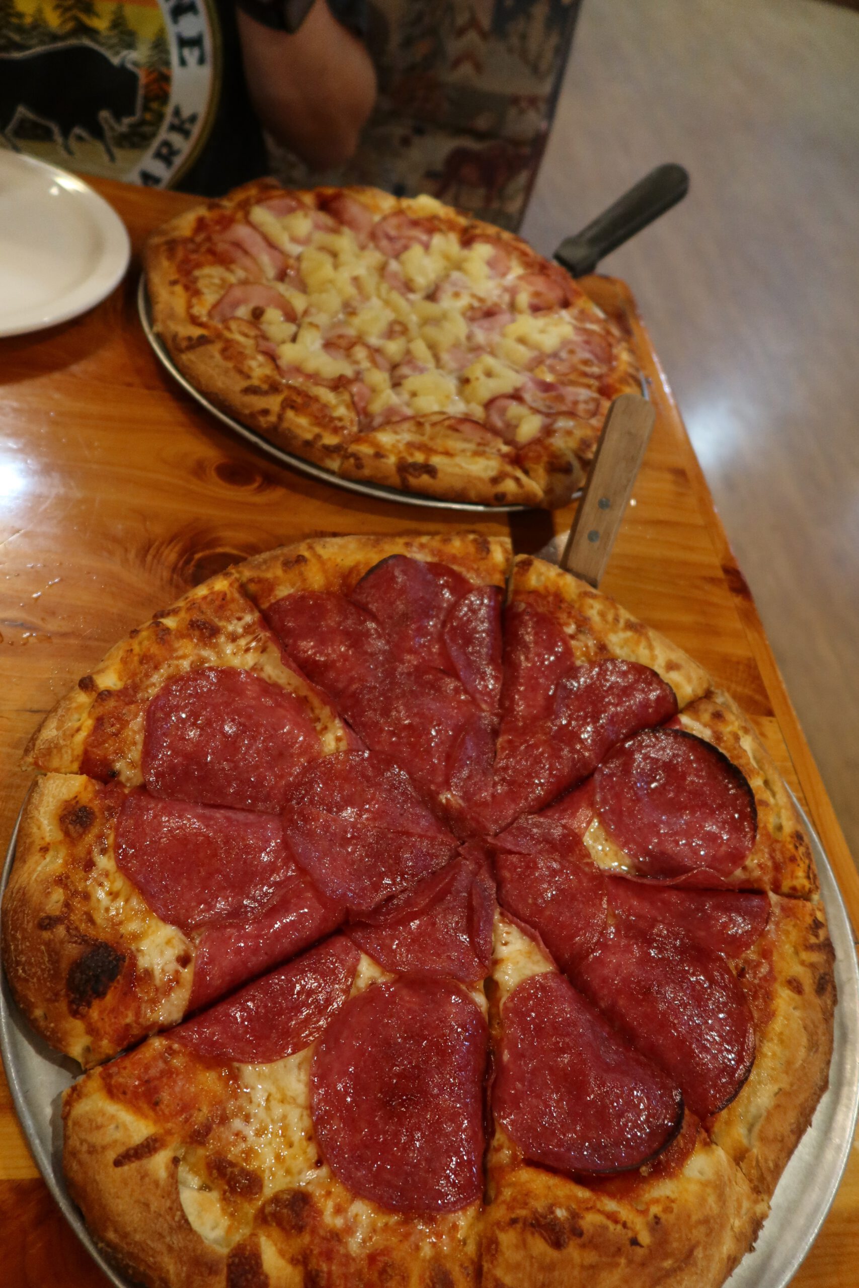 Large freshly baked pizzas at Outpost Pizza Pub & Grill John Day, featuring a classic pepperoni pizza in the foreground and a ham and pineapple pizza in the background, served on metal pizza trays on a wooden table, highlighting the generous portions and classic American pizza style in John Day, Oregon.