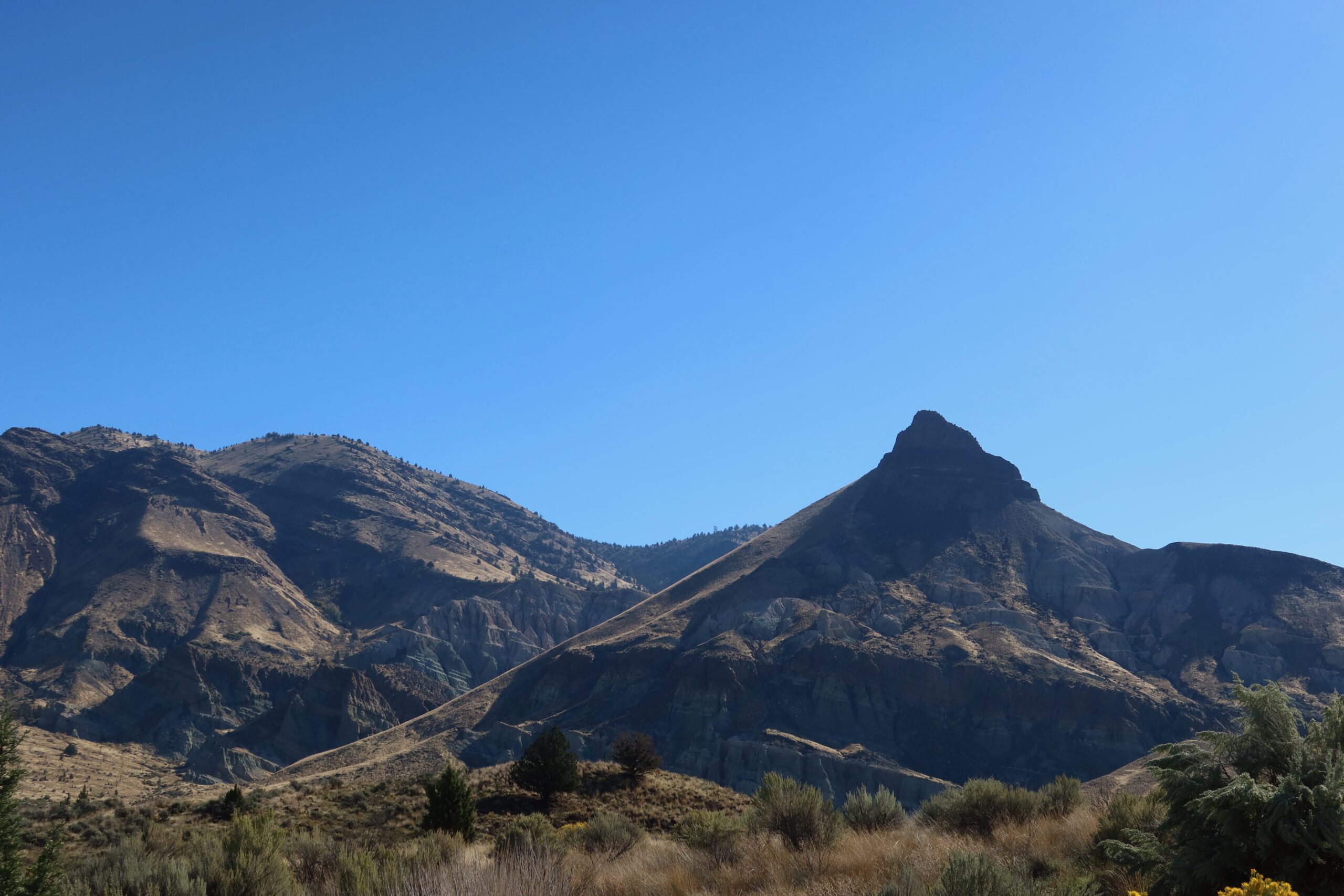 Sharp triangular rock peak rising from layered volcanic hills in eastern Oregon, surrounded by dry grassland and rugged badlands under a clear blue sky near the John Day Fossil Beds.