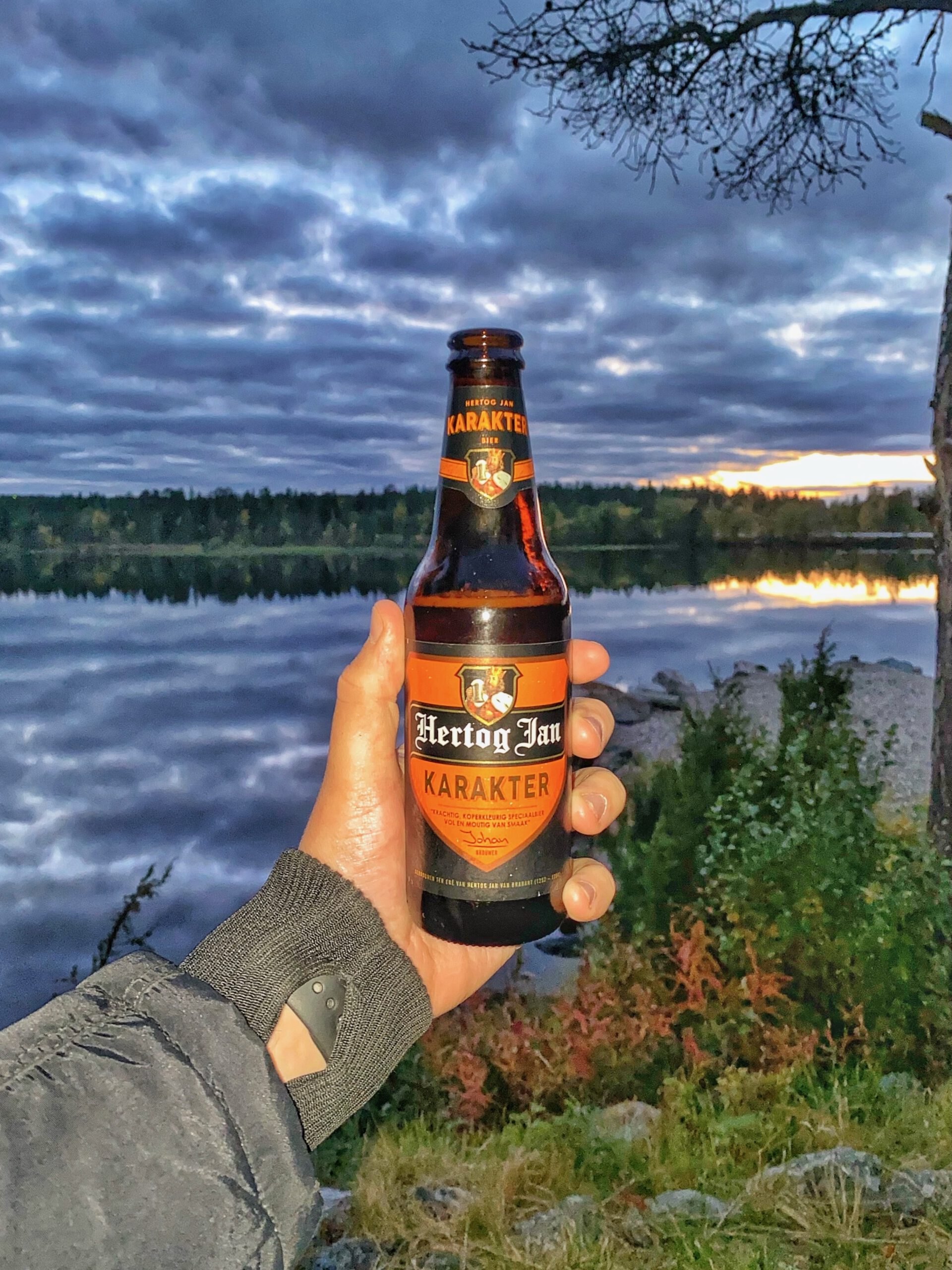 Hertog Jan Karakter speciaalbier van Hertog Jan Brouwerij, vastgehouden bij een rustig meer tijdens zonsondergang bij Hetan Kota in Enontekiö, Finland, omringd door Laplandse natuur en herfstkleuren.
