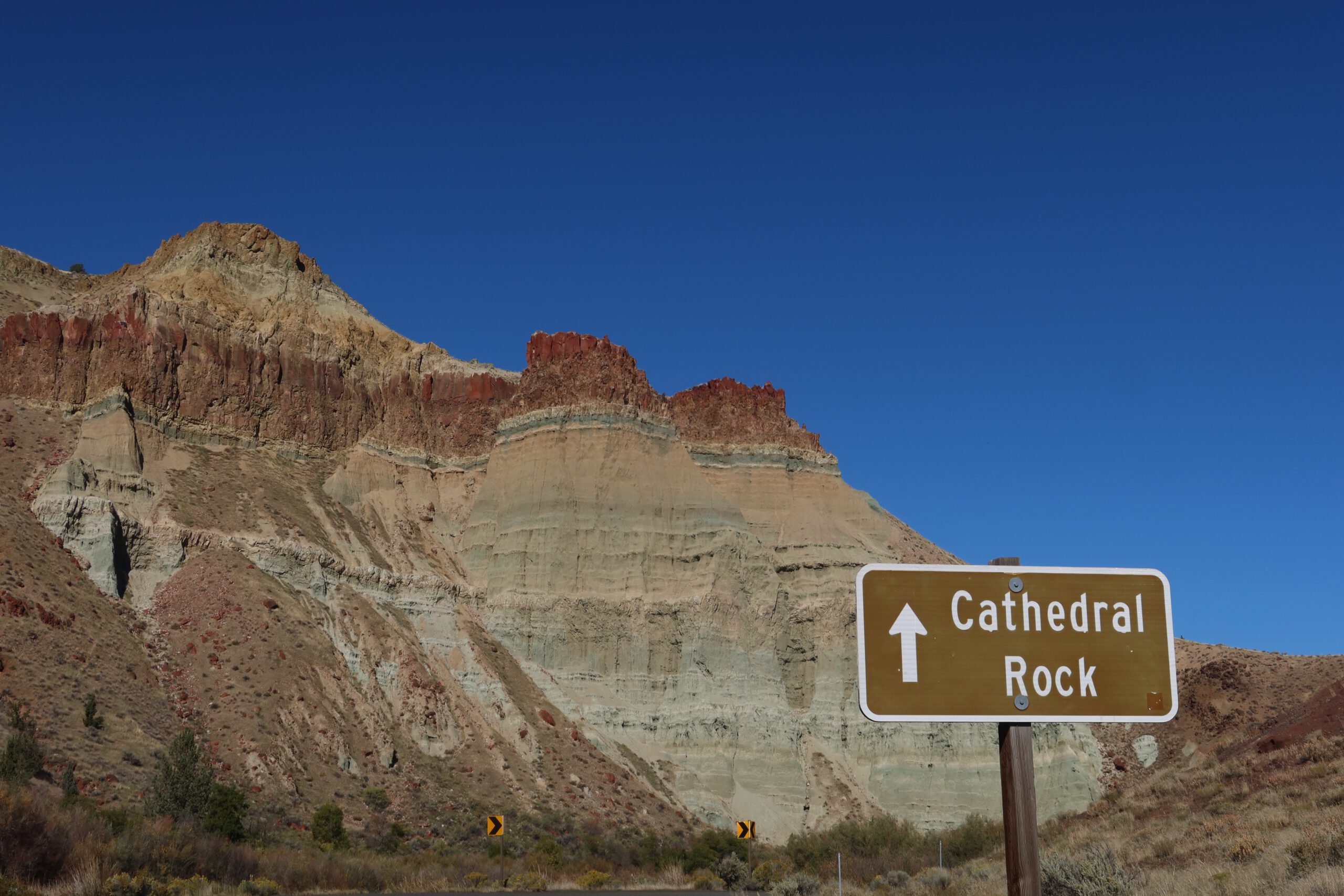 Cathedral Rock sign pointing toward layered cliffs at John Day Fossil Beds National Monument, highlighting colorful volcanic rock formations under a clear blue sky in Eastern Oregon