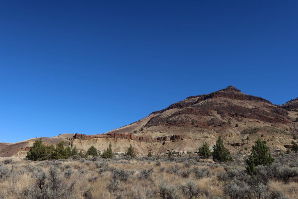 Wide landscape view of layered hills and badlands within John Day Fossil Beds National Monument, featuring sagebrush plains and volcanic rock formations under a clear blue sky