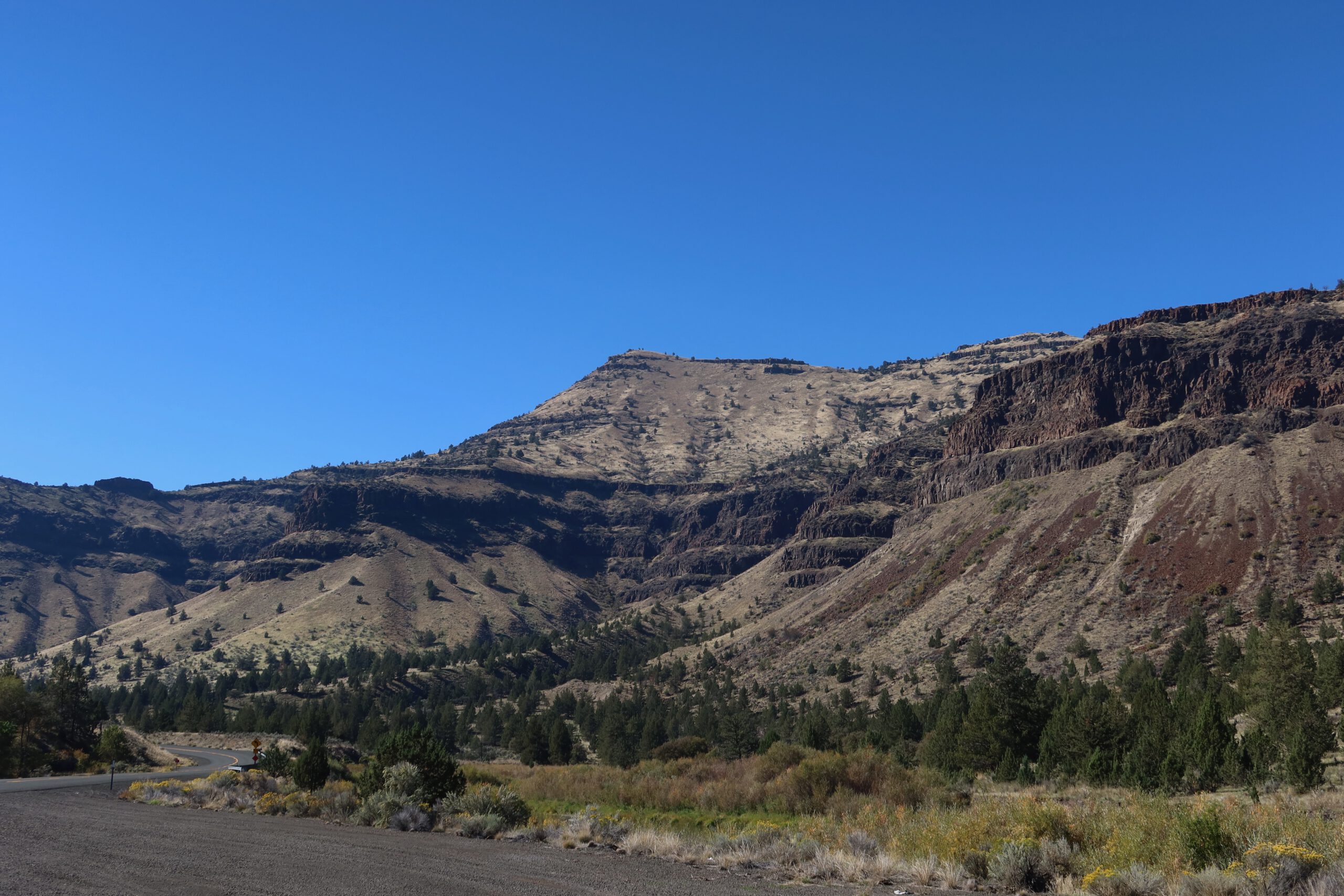 Layered volcanic cliffs and rugged badlands landscape in eastern Oregon under a clear blue sky, showcasing dramatic rock formations near the John Day Fossil Beds.
