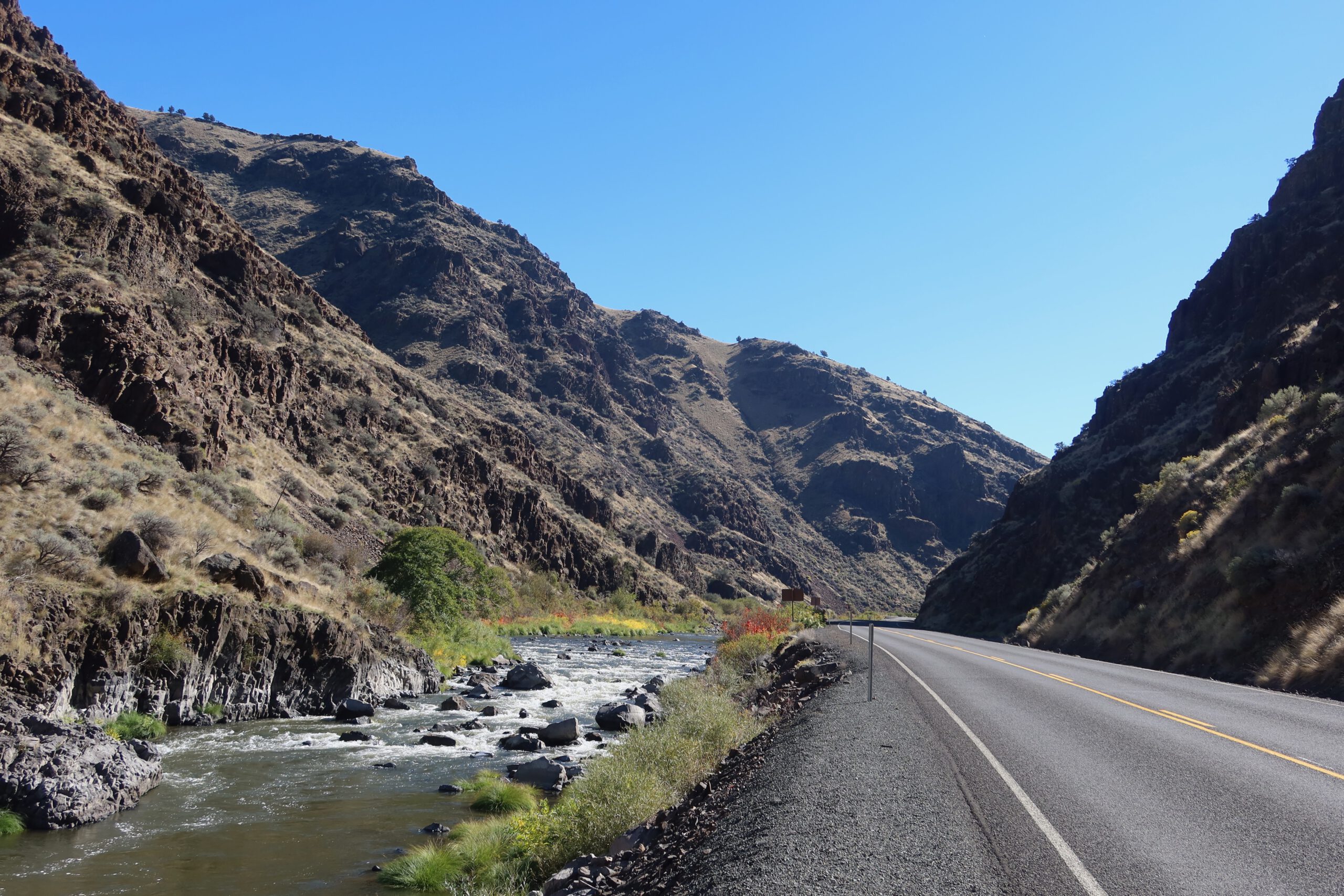 Scenic roadside view along Highway 19 near John Day Fossil Beds National Monument, with a winding river flowing through a rocky canyon and steep Oregon hills under a clear blue sky.