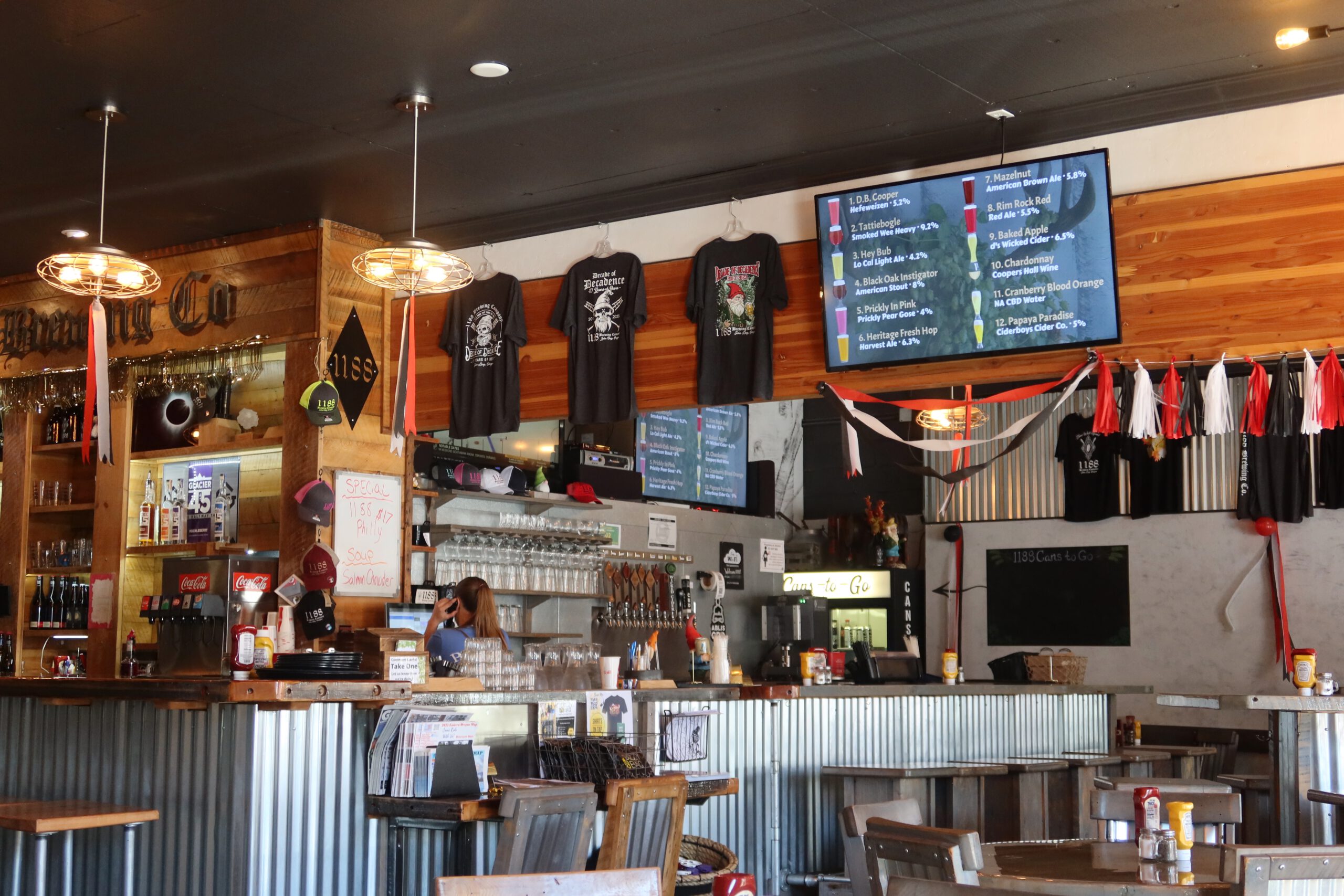 Interior of 1188 Brewing Company in John Day Oregon, showing the rustic bar area with beer taps, merchandise, wooden decor, and digital tap list screens displaying local craft beers in a small town brewery atmosphere.