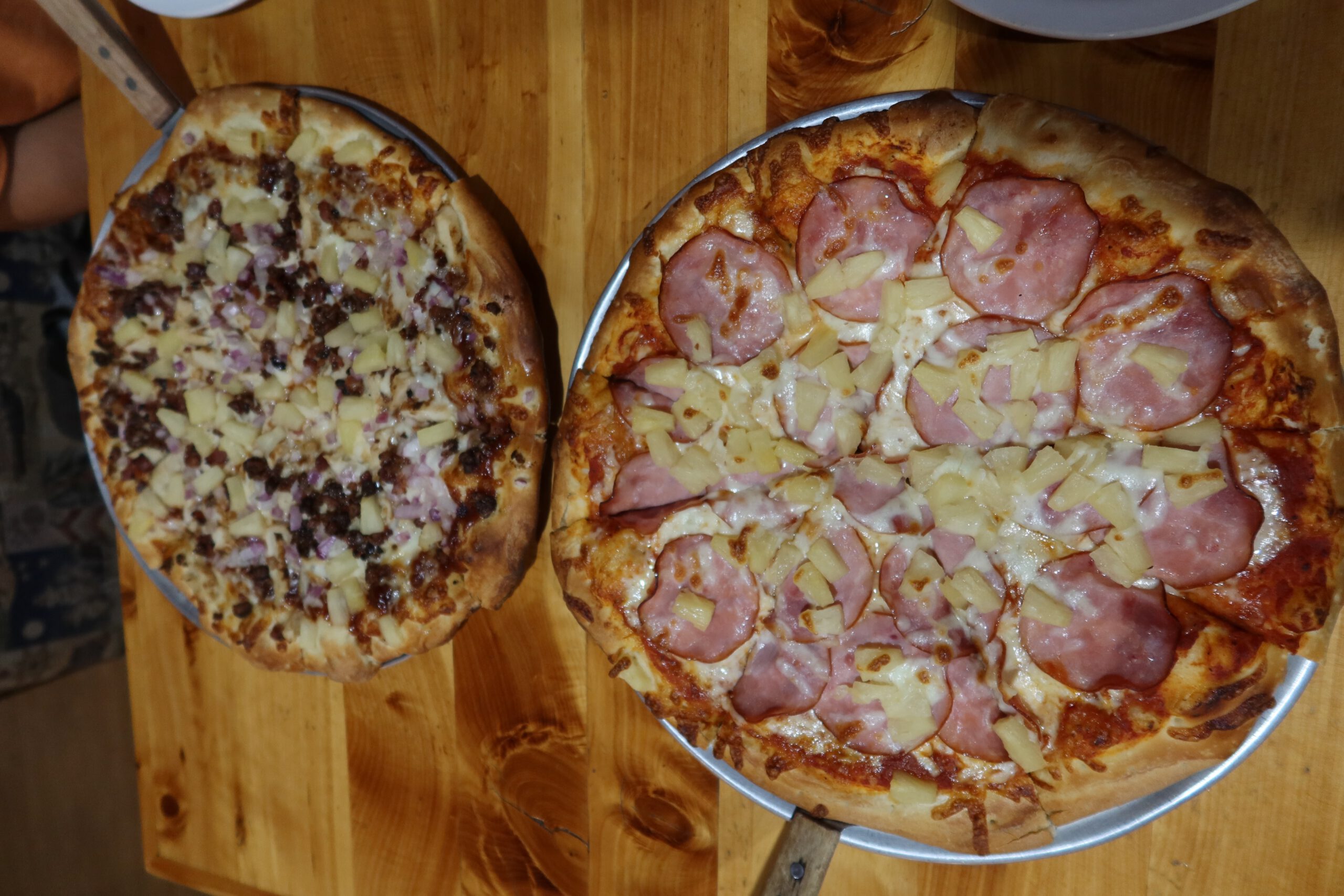 Overhead view of two freshly baked pizzas at Outpost Pizza Pub & Grill John Day, featuring a ham and pineapple pizza and a beef and onion pizza served on metal trays on a wooden table, highlighting generous portions and classic American pizza styles in John Day, Oregon.