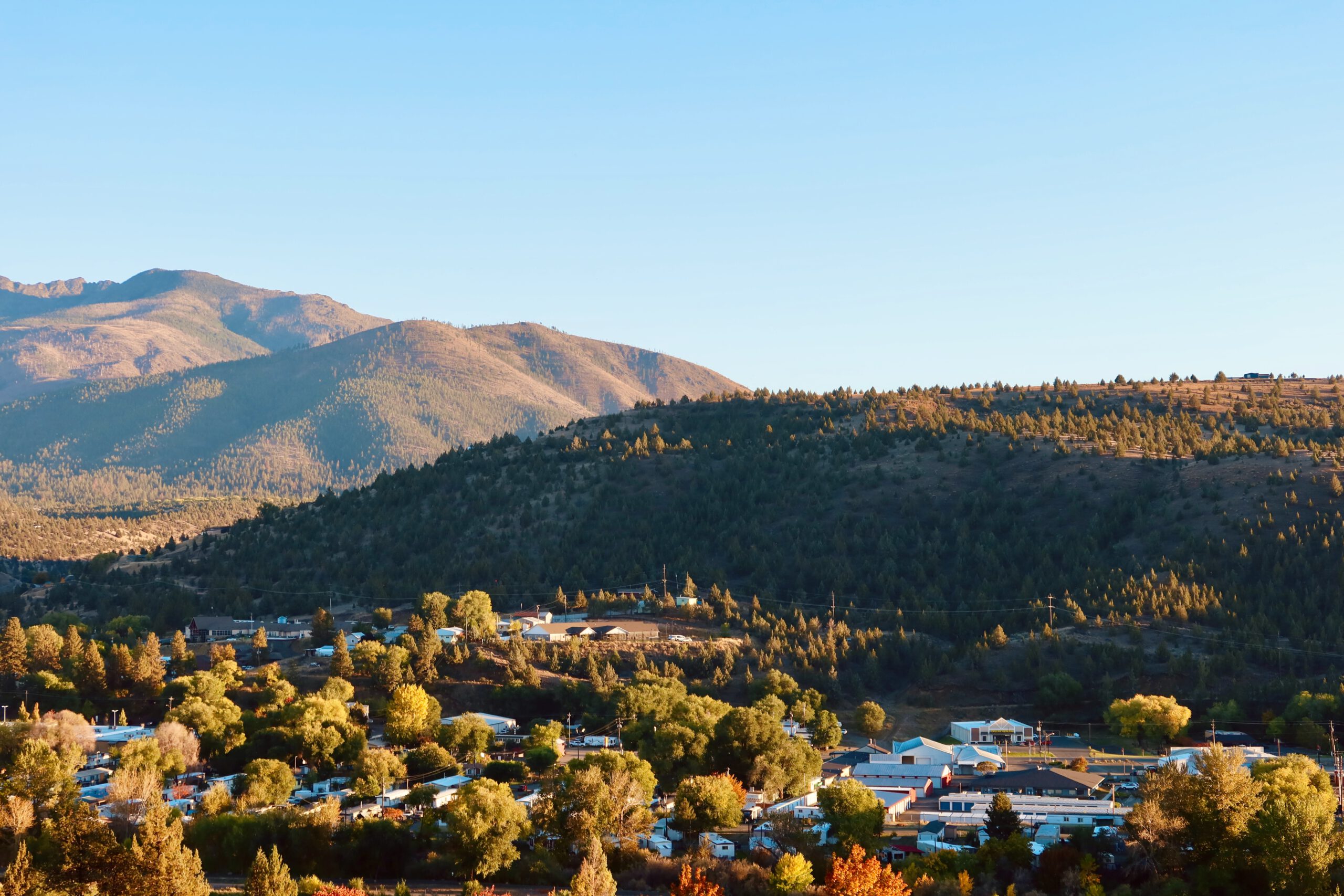 Panoramic view of John Day, Oregon, taken from an elevated viewpoint, showing the small town surrounded by rolling hills, pine covered mountains, and warm evening light under a clear blue sky.