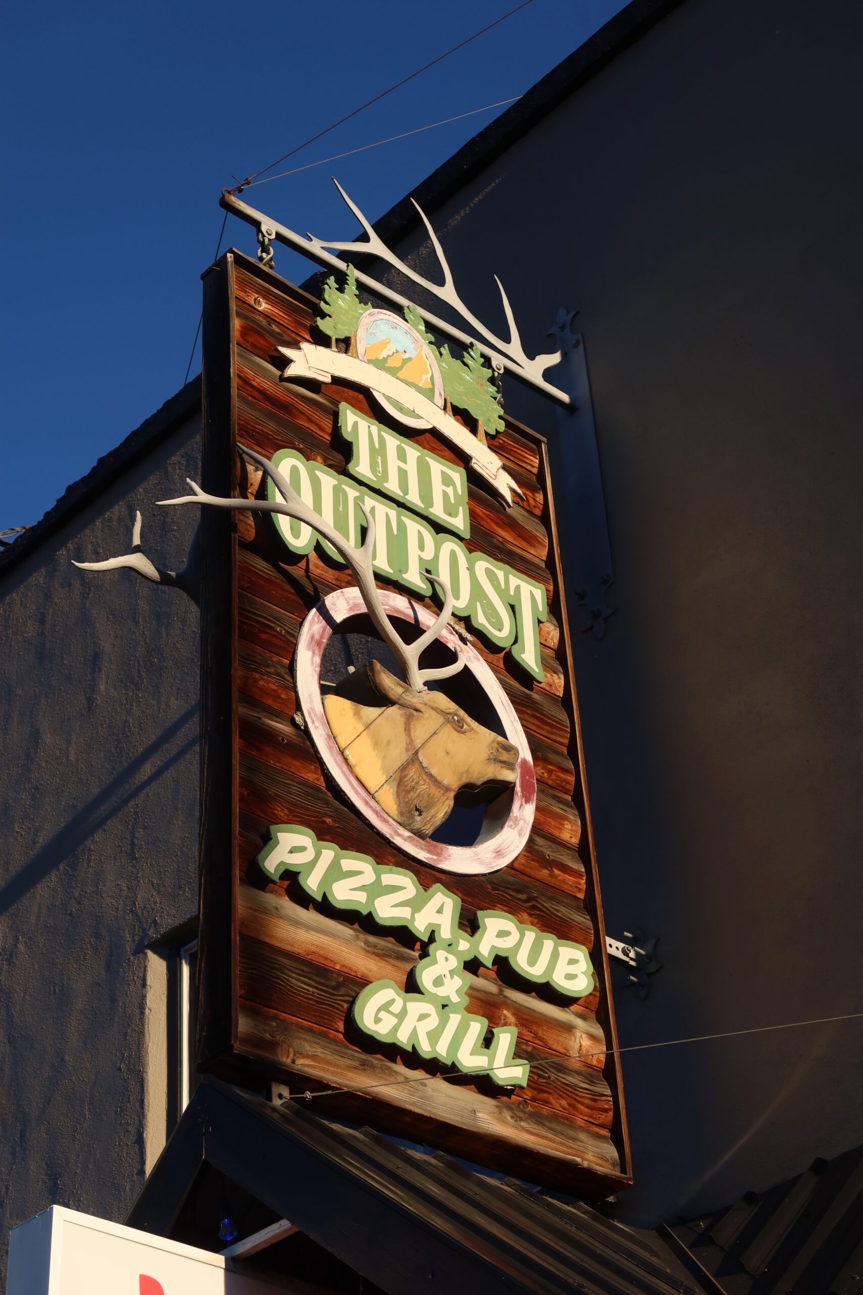 Exterior wooden sign of Outpost Pizza Pub & Grill John Day, featuring a rustic elk head logo and antler design, mounted on the building facade under a clear blue sky, showcasing the classic small town American pizza pub atmosphere in John Day, Oregon.