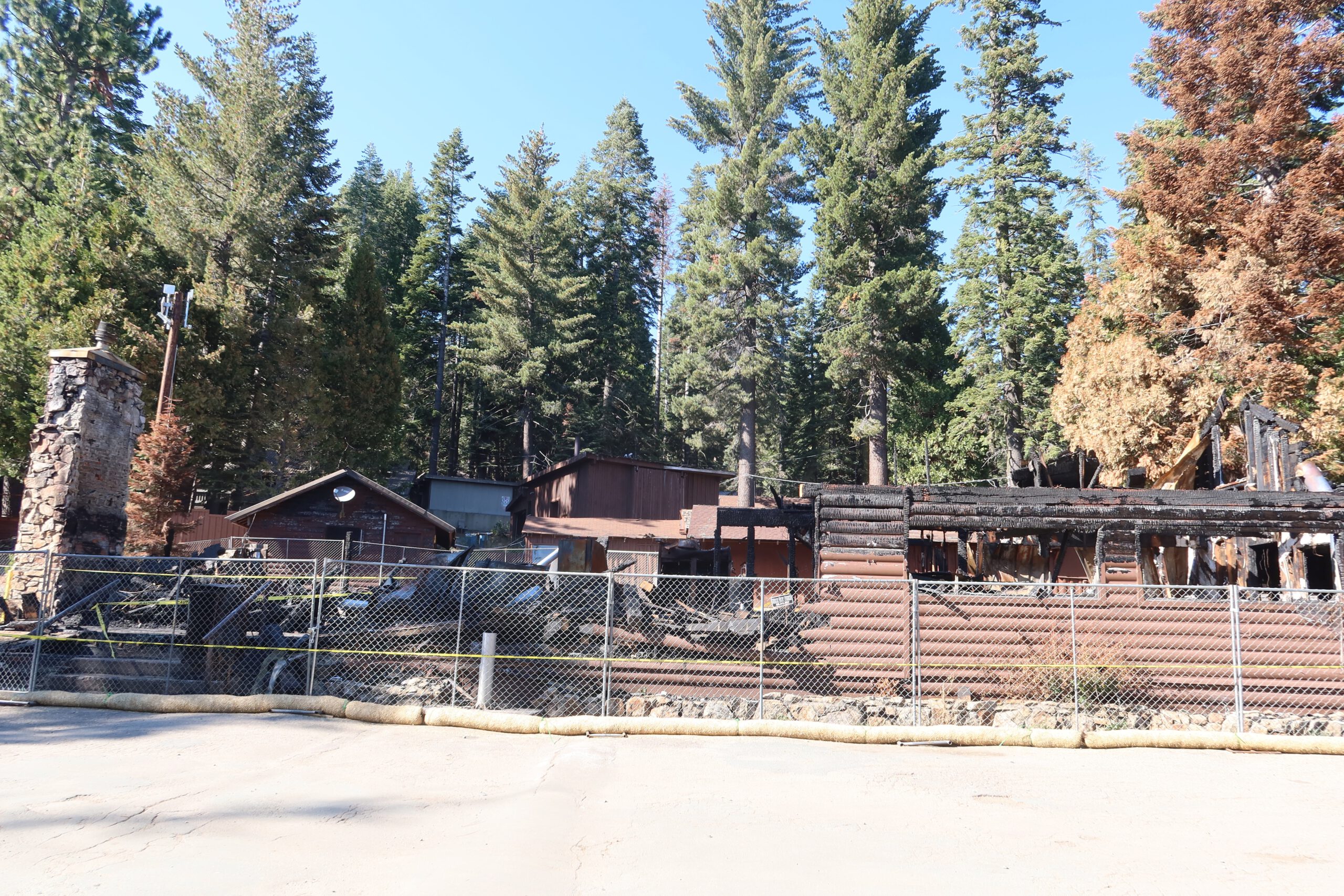 Wide view of Bear River Lake Resort after the 2023 fire in Amador County, California, showing the burned lodge site behind safety fencing and surrounding pine forest