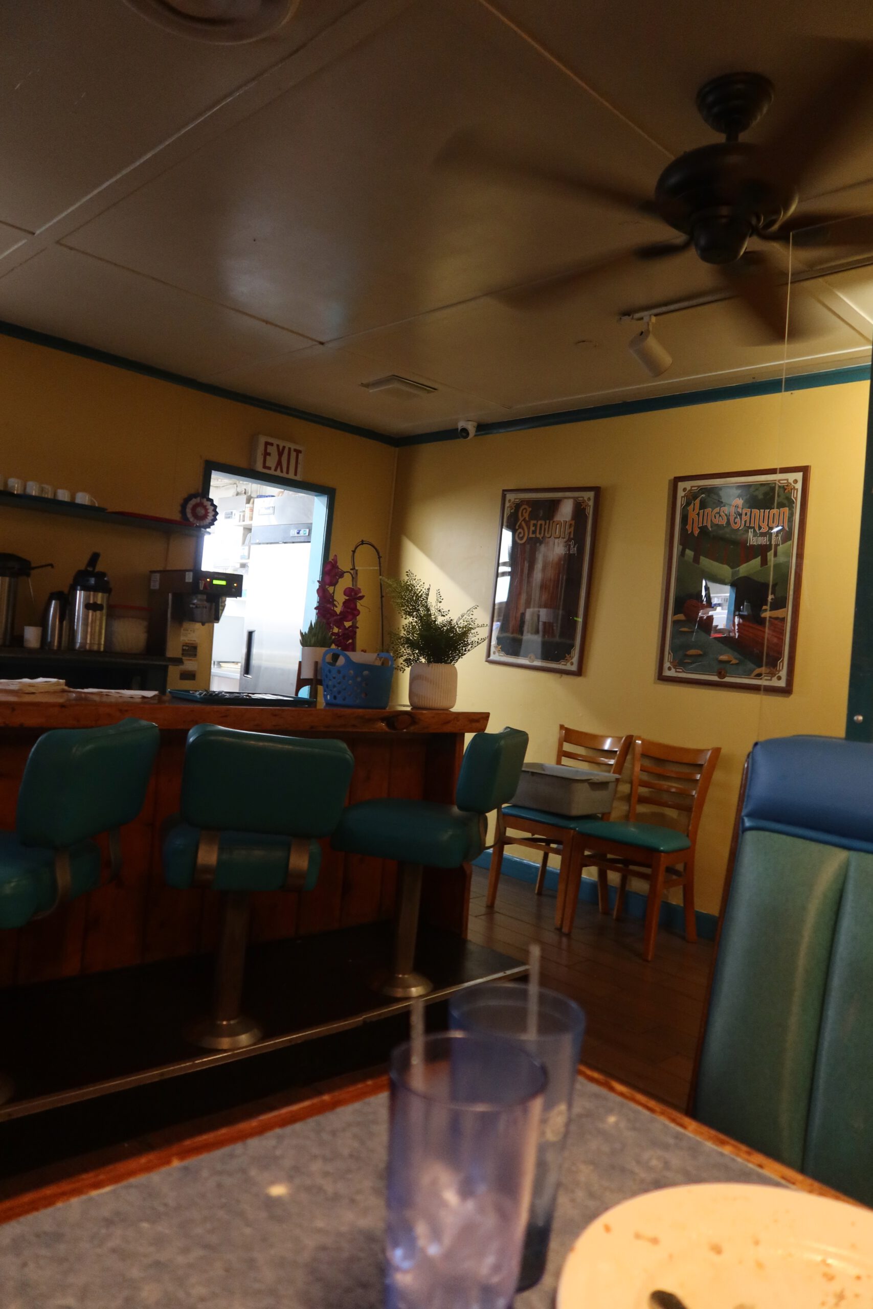 Interior view of Lucky Buck Cafe California showing teal bar stools, wooden chairs, ceiling fan, framed National Park posters, and a cozy classic American diner atmosphere near Yosemite.