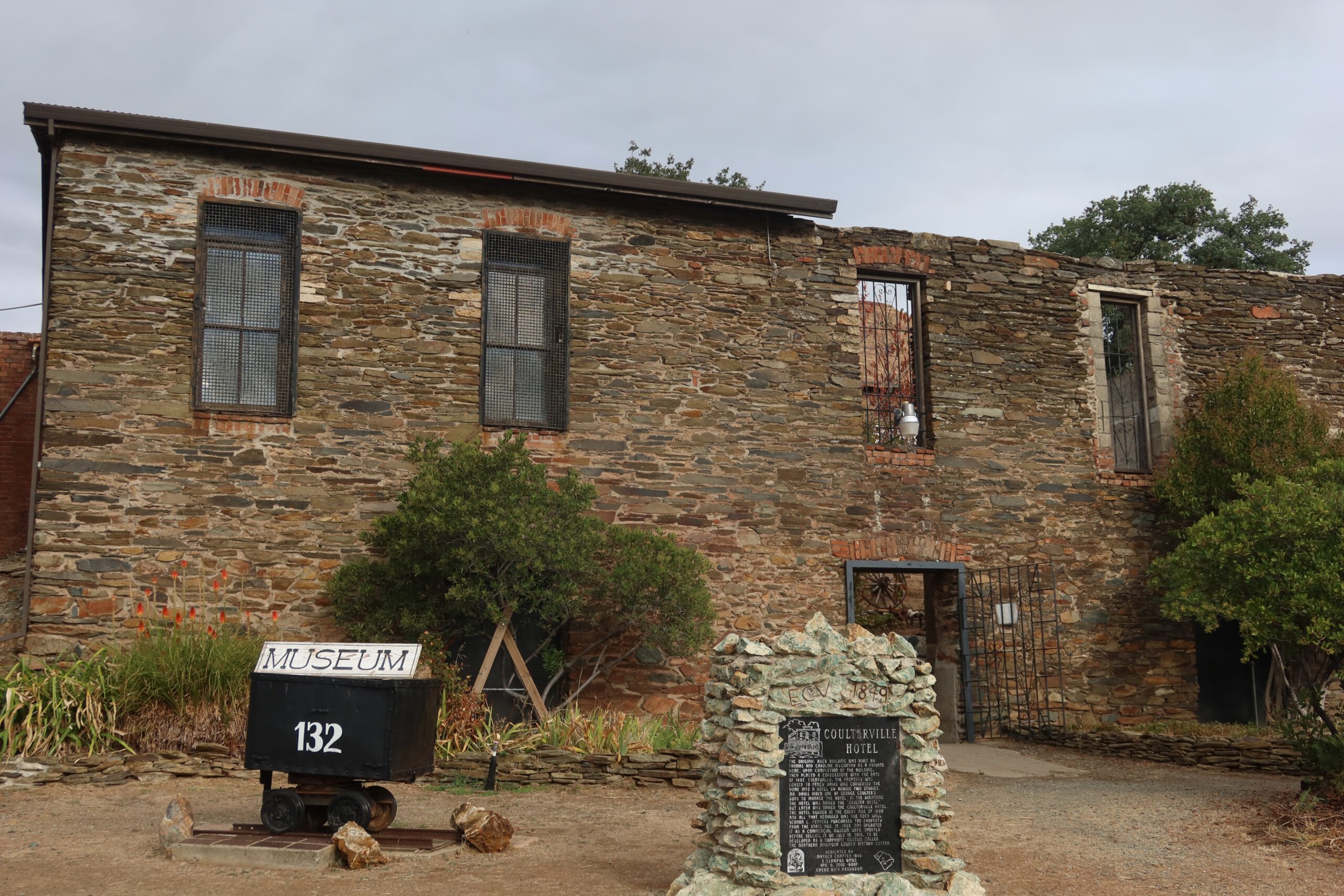 Stone museum building at the Coulterville History Center in Coulterville, California, featuring historic gold rush architecture, a mine cart museum sign, and the Coulterville Hotel historical marker along CA 49
