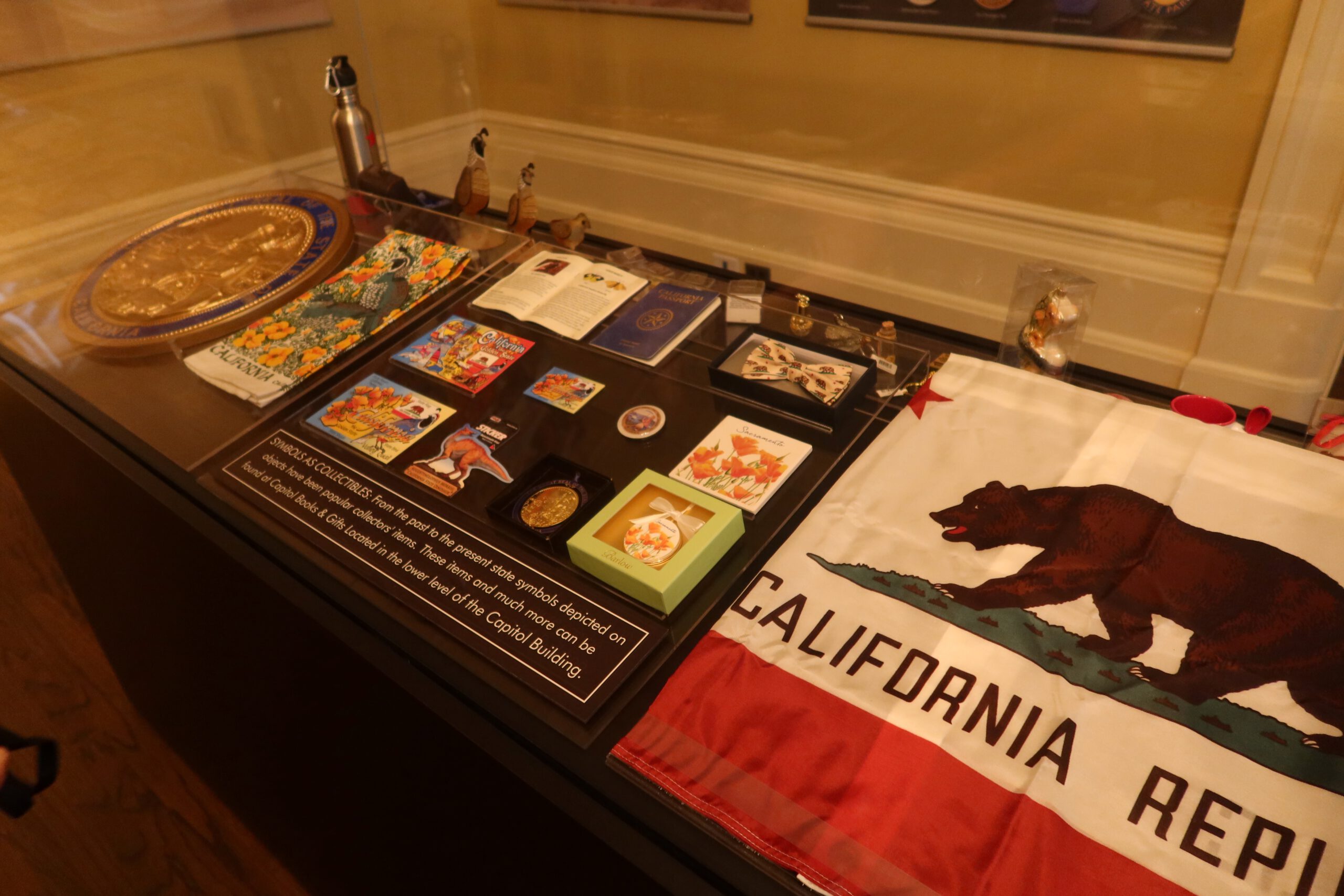 Museum display case inside the California State Capitol Museum, showing historic California symbols such as the Bear Flag, books, postcards, medals, and cultural artifacts explaining state identity and Gold Rush era heritage.