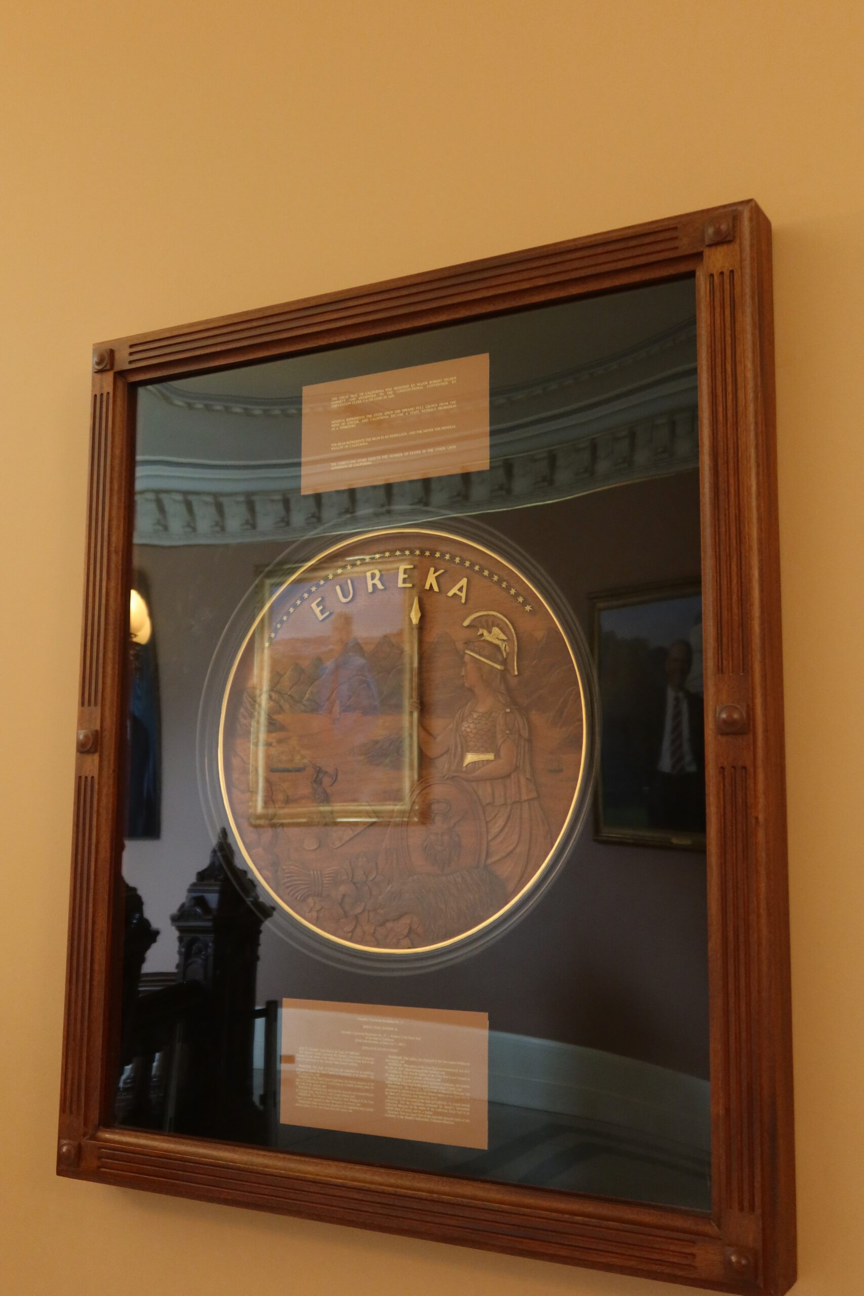 Framed Eureka state seal display inside the California State Capitol Museum, showing the official California emblem with Minerva, mountains, and Gold Rush symbolism mounted on a historic interior wall.