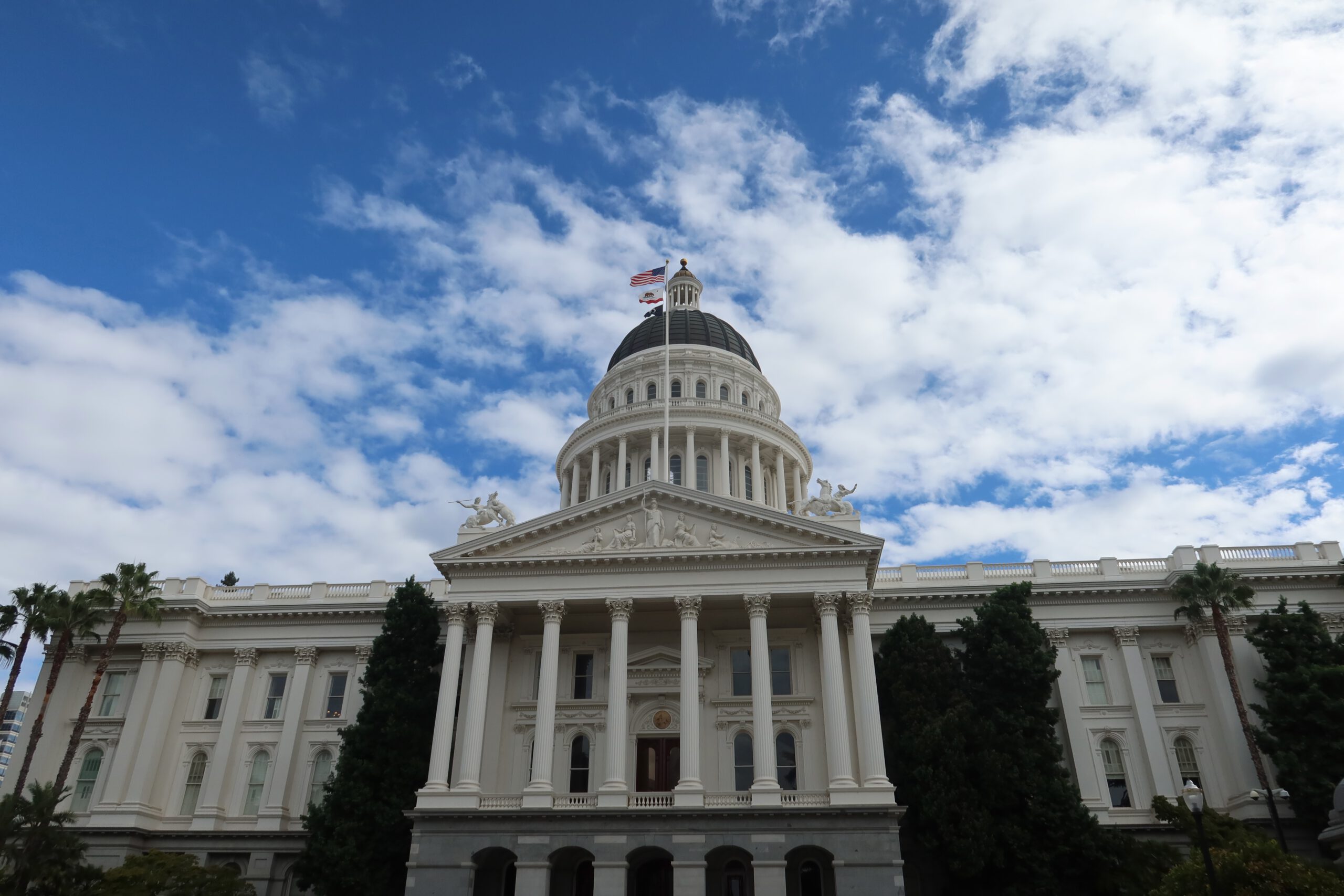 California State Capitol building in Sacramento, featuring the iconic white neoclassical facade, tall Corinthian columns, and the central dome with the United States and California flags under a blue sky.