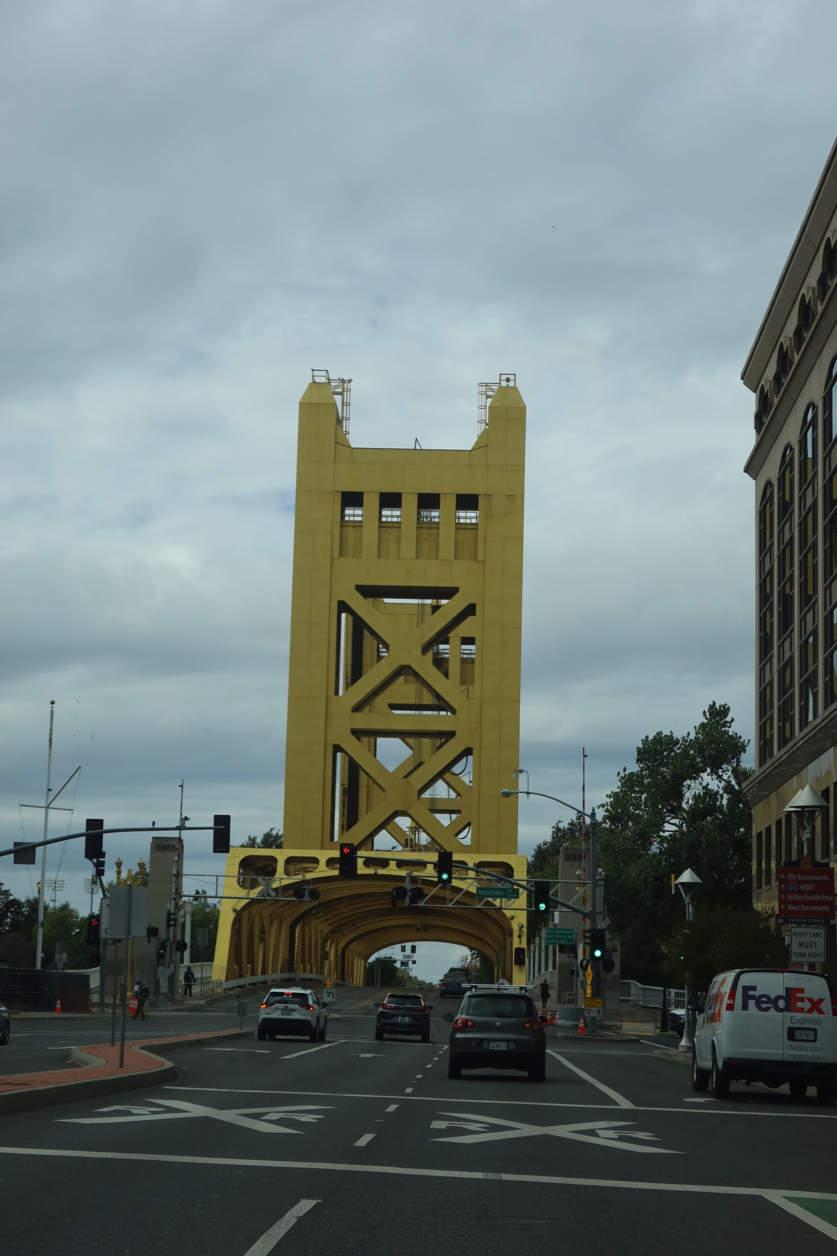 Driving toward downtown Sacramento with a clear view of the iconic yellow Tower Bridge, a major landmark near the California State Capitol Museum, with cars crossing the bridge under an overcast sky.