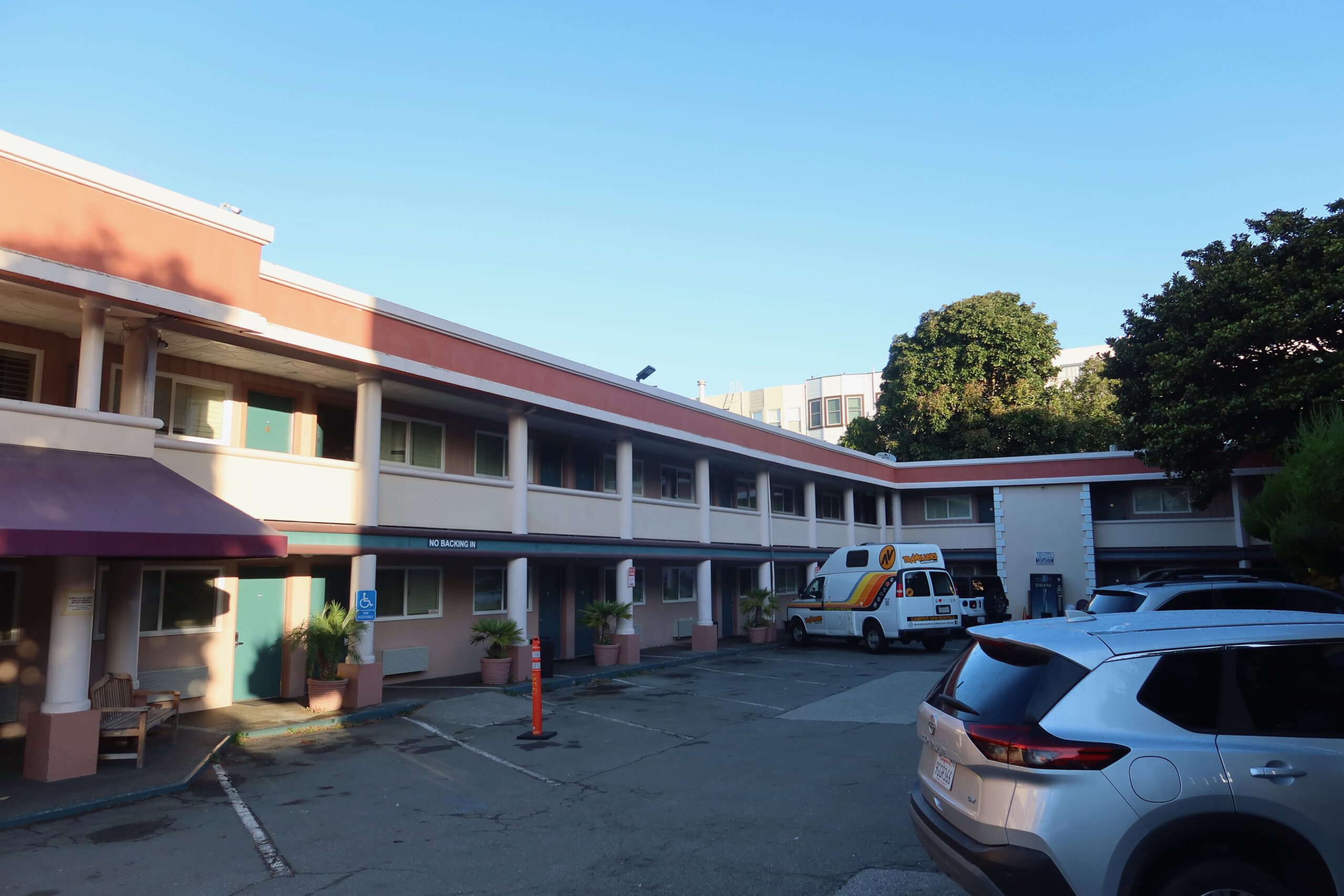 Wide courtyard view of the Town House Motel San Francisco, featuring pastel pink architecture, classic motel balconies, parked cars, and a clear blue sky.