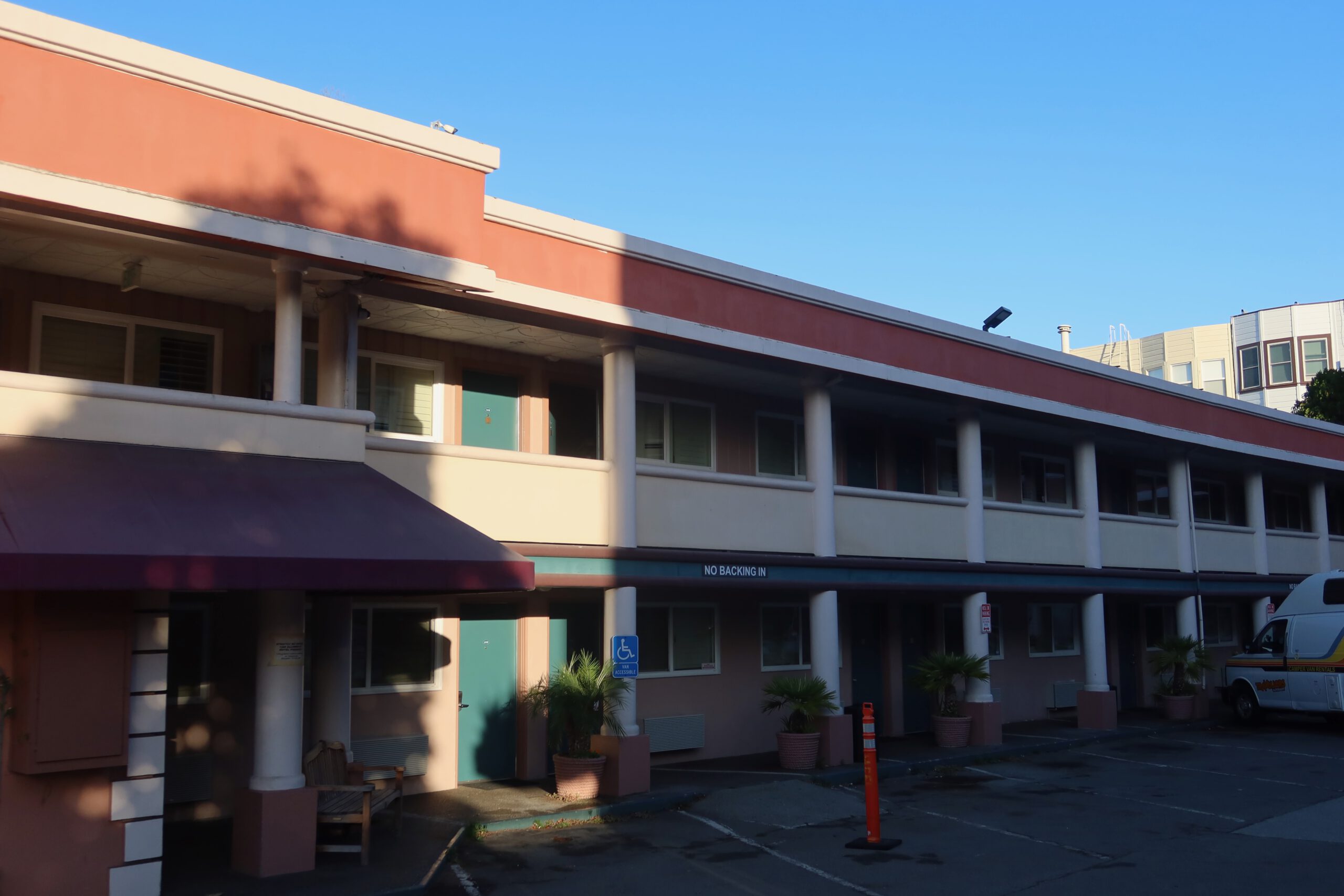 Exterior view of the Town House Motel in San Francisco, showing the two-story mid-century motel building with teal room doors, covered walkways, and parking area in early morning light.