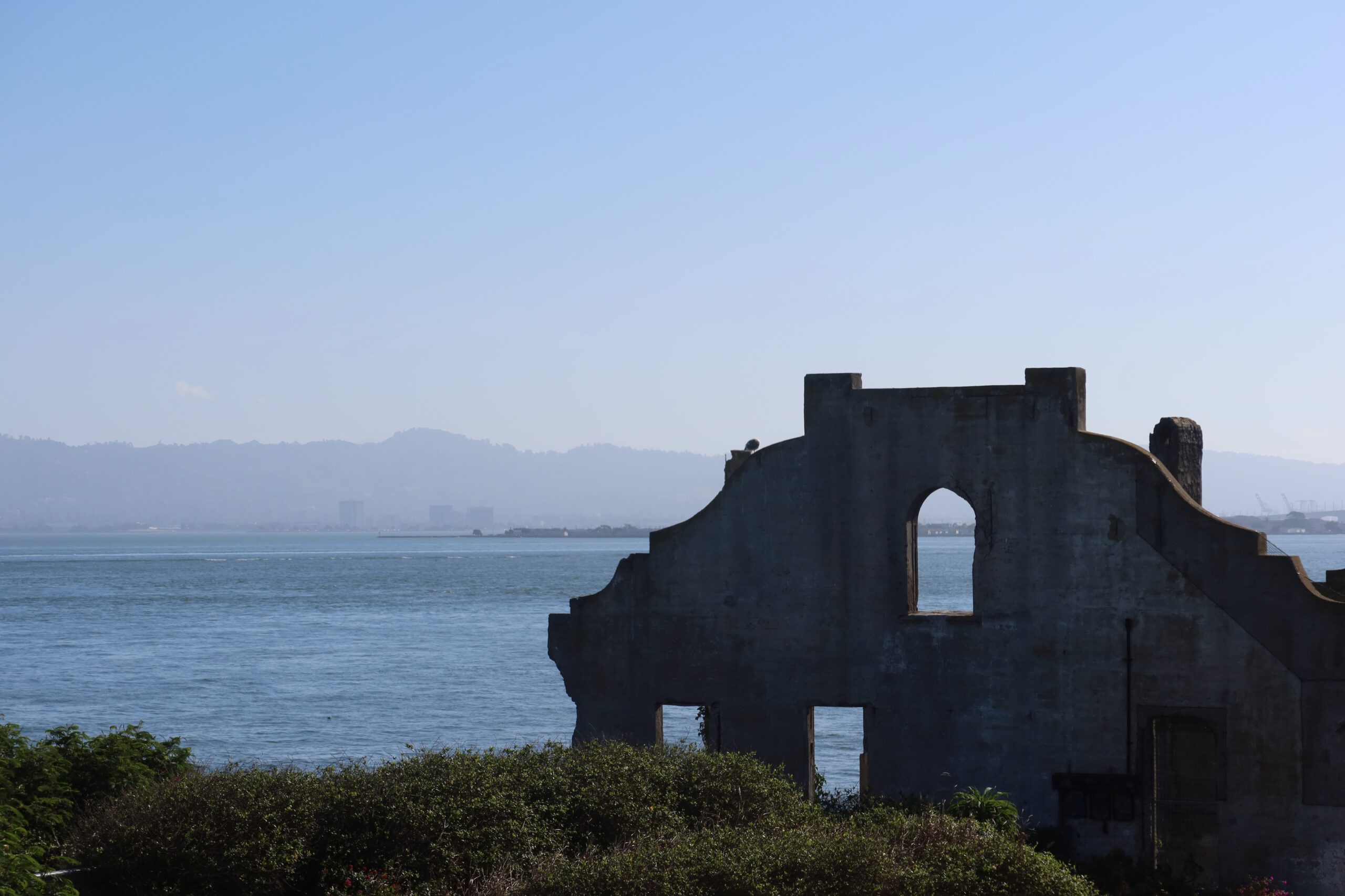 Concrete ruins on Alcatraz Island with a large arched window opening, overlooking San Francisco Bay and distant hills