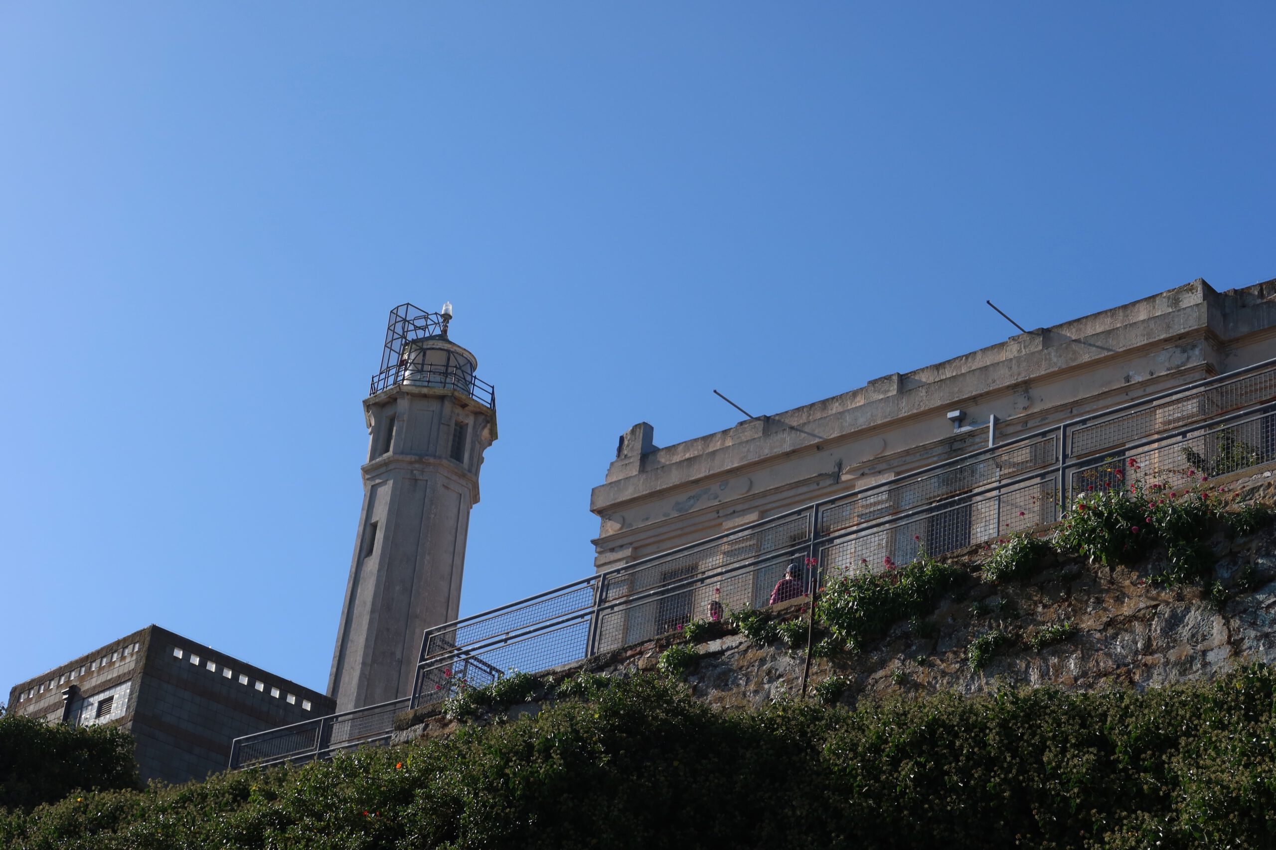 Alcatraz Island lighthouse and historic prison buildings seen from below, surrounded by greenery under a clear blue sky