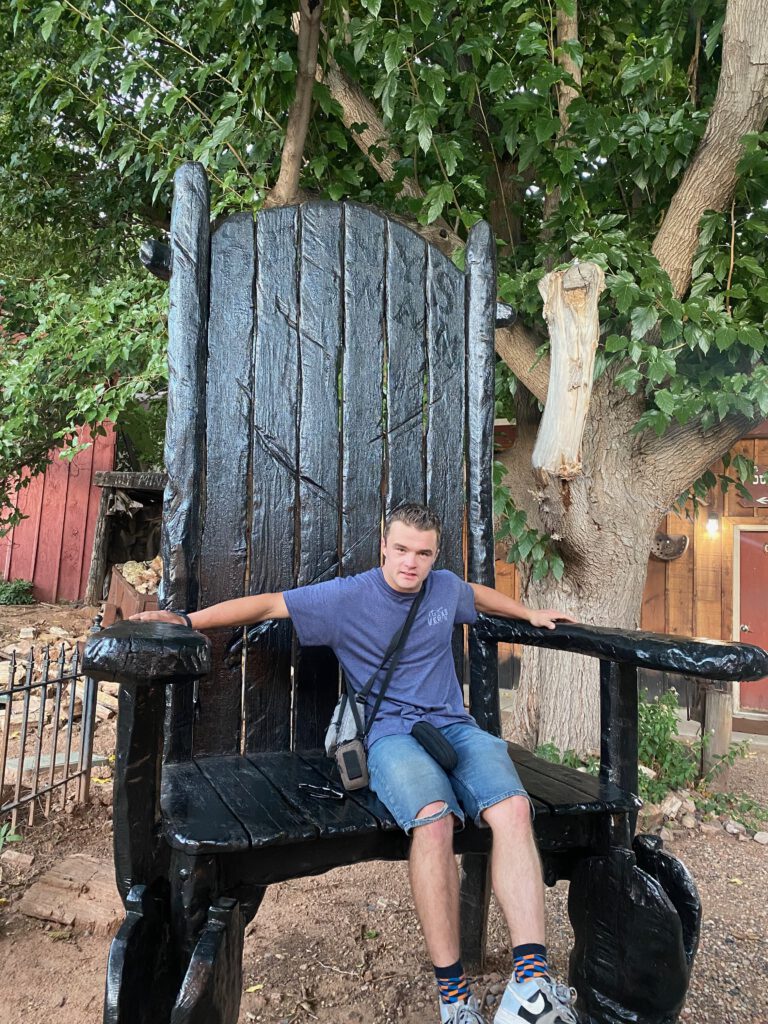 Visitor sitting on an oversized wooden chair at Iron Horse Kanab Utah, surrounded by trees and western style buildings, highlighting the quirky outdoor atmosphere.