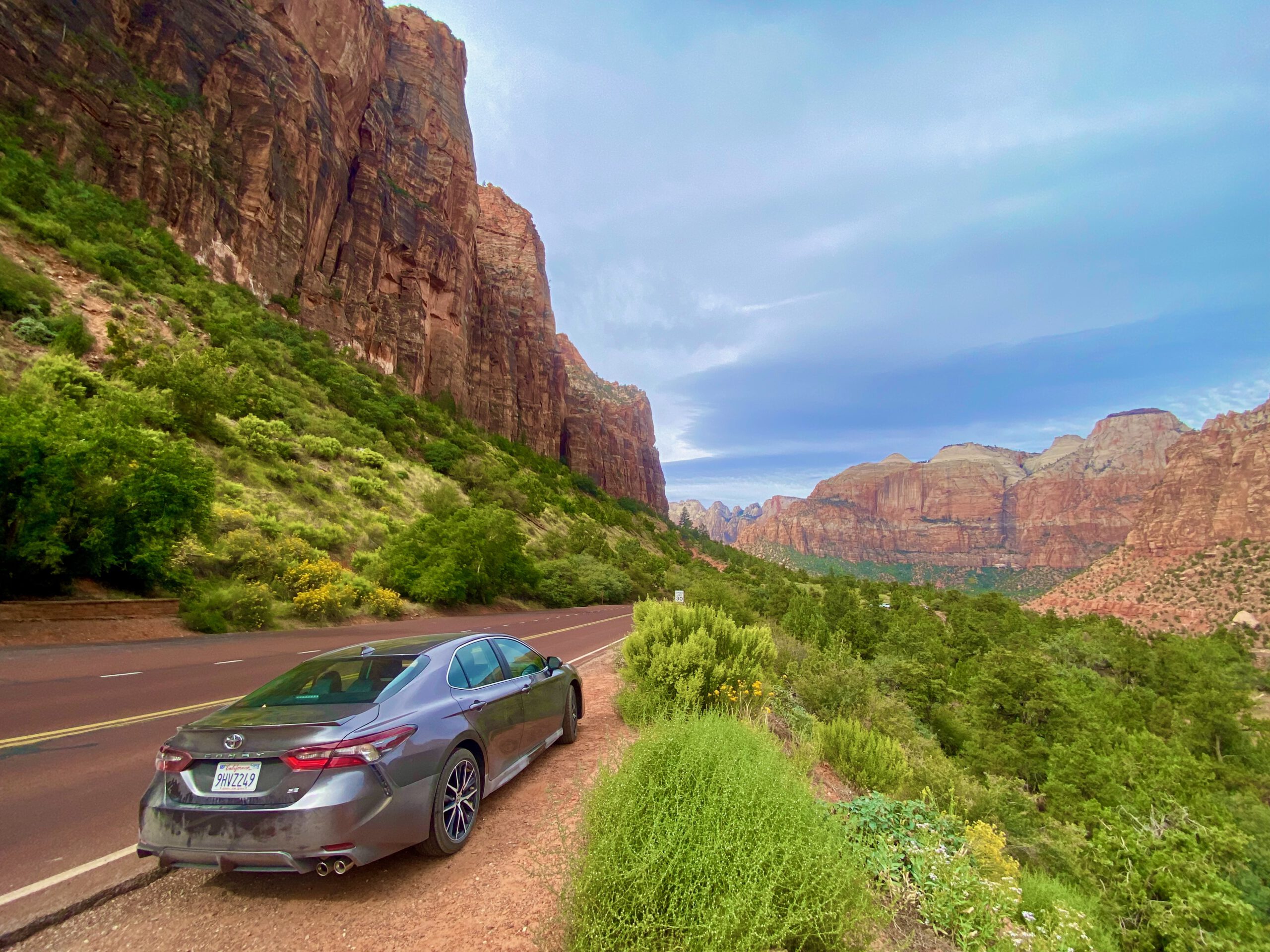 Parked car along Zion Canyon Scenic Drive with towering canyon walls, green vegetation, and dramatic blue sky in Zion National Park