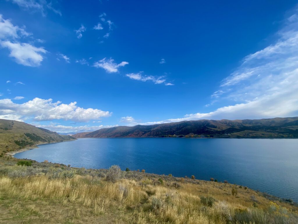 Wide scenic view of Echo Reservoir near Coalville Utah, with bright blue water, golden grasses in the foreground, and rolling hills under a vast blue sky, photographed during a Utah road trip stop.