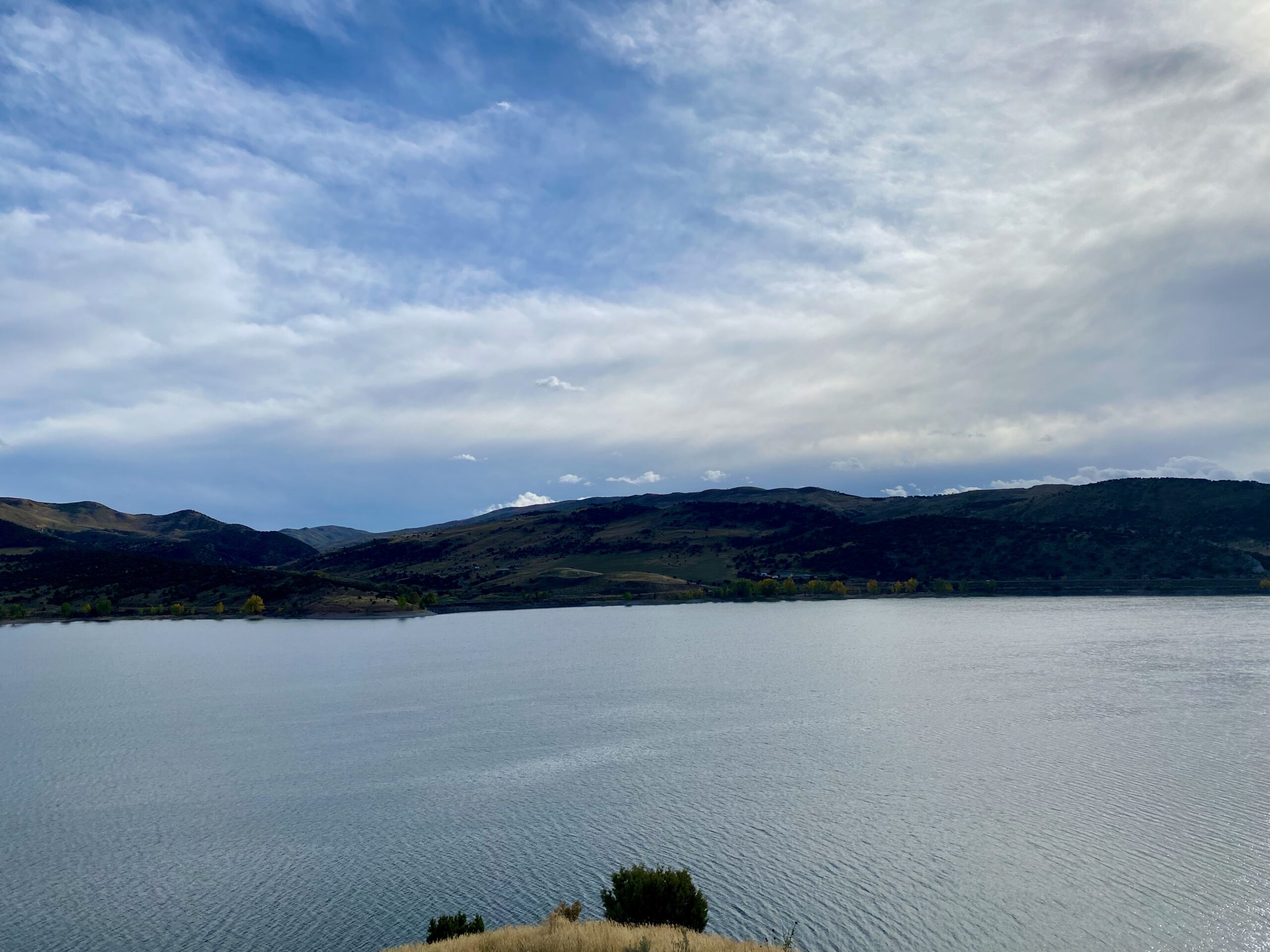 Calm view across Echo Reservoir near Coalville Utah, with still blue water reflecting soft clouds and dark rolling hills in the distance, captured during a quiet Utah road trip stop.