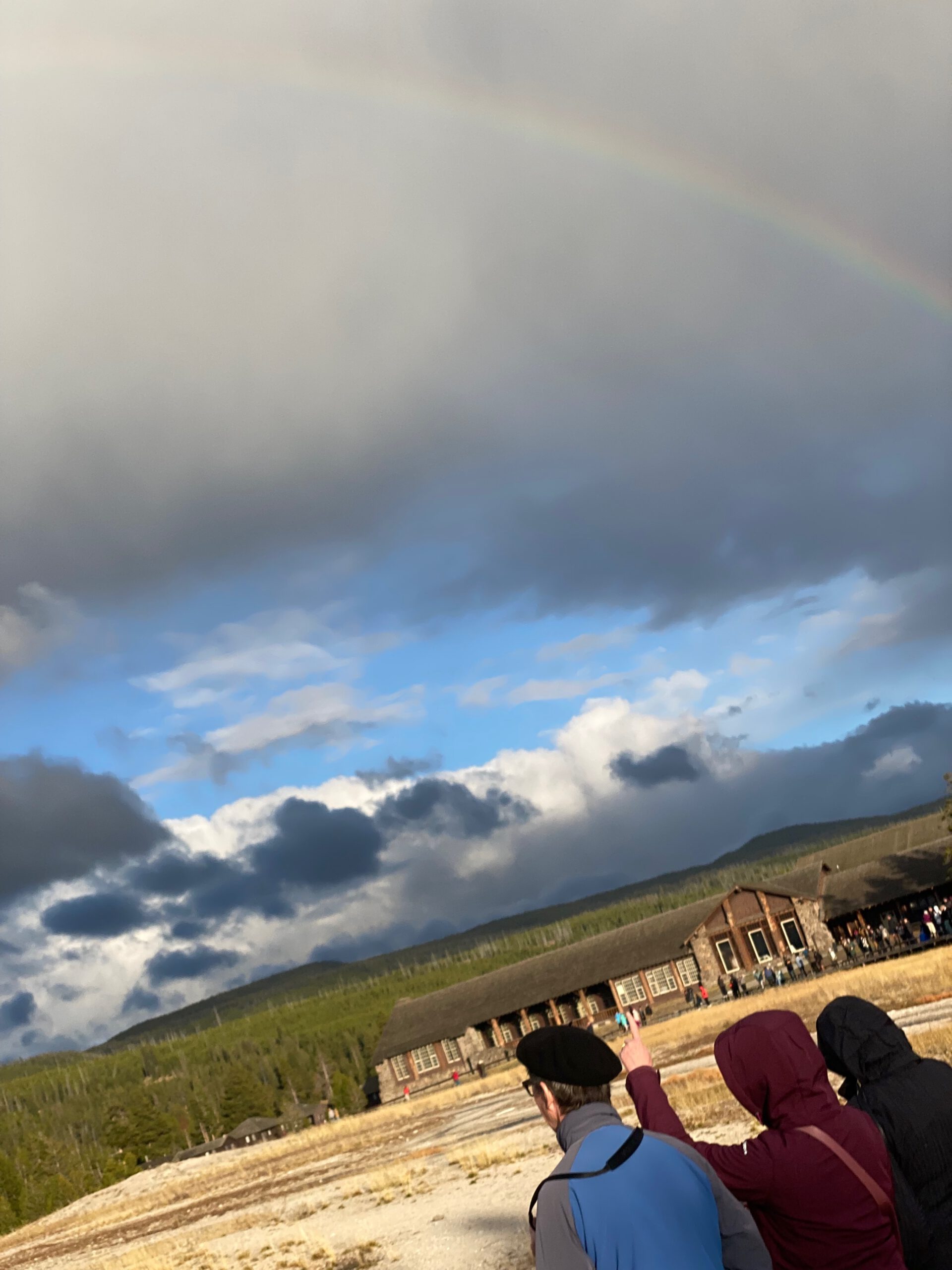 Visitors watching Old Faithful area under dramatic clouds and a faint rainbow in Yellowstone National Park, Wyoming