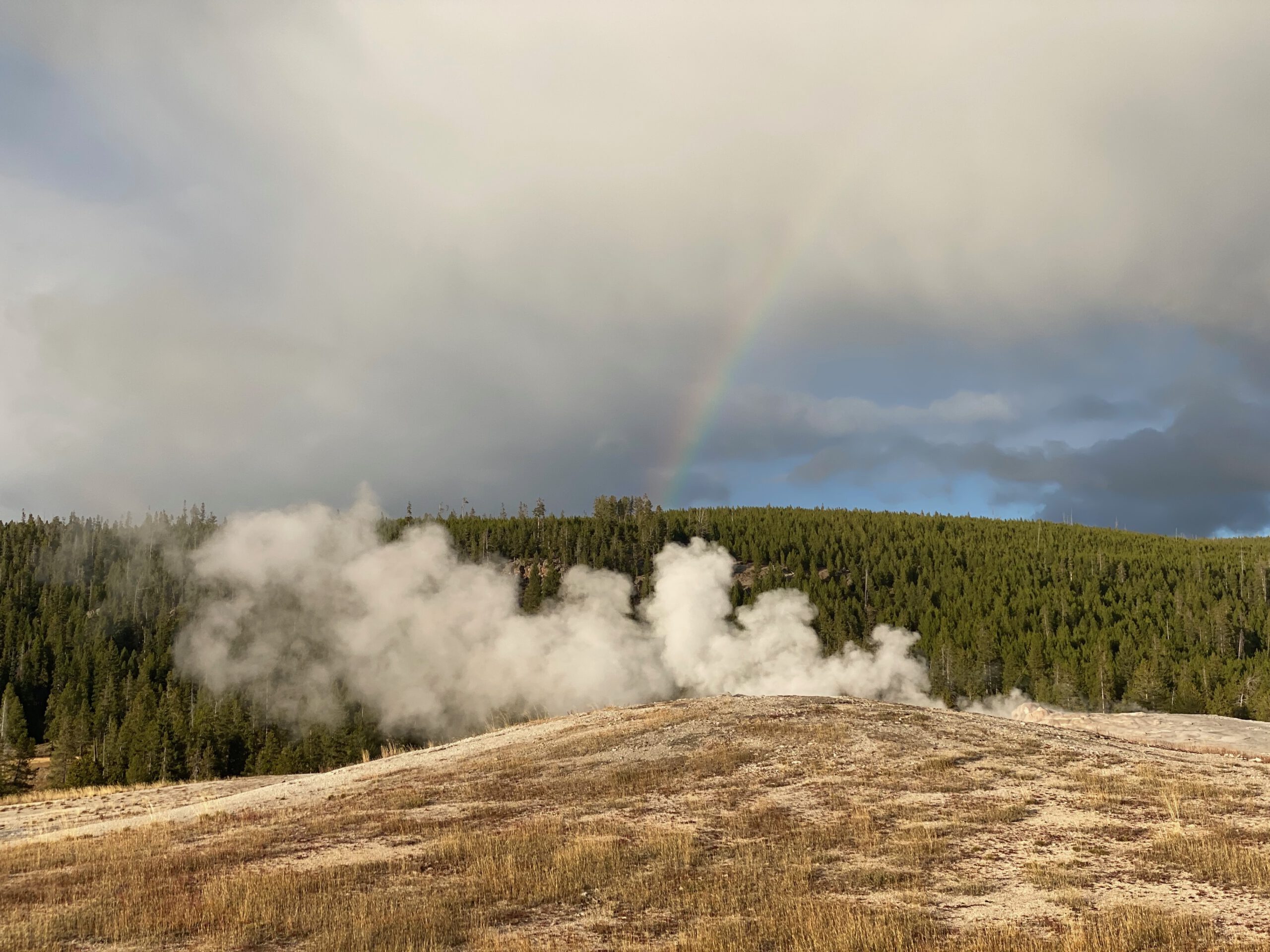 Steam rising from multiple geysers in Yellowstone National Park with a faint rainbow in the sky, showing intense geothermal activity beneath the surface.