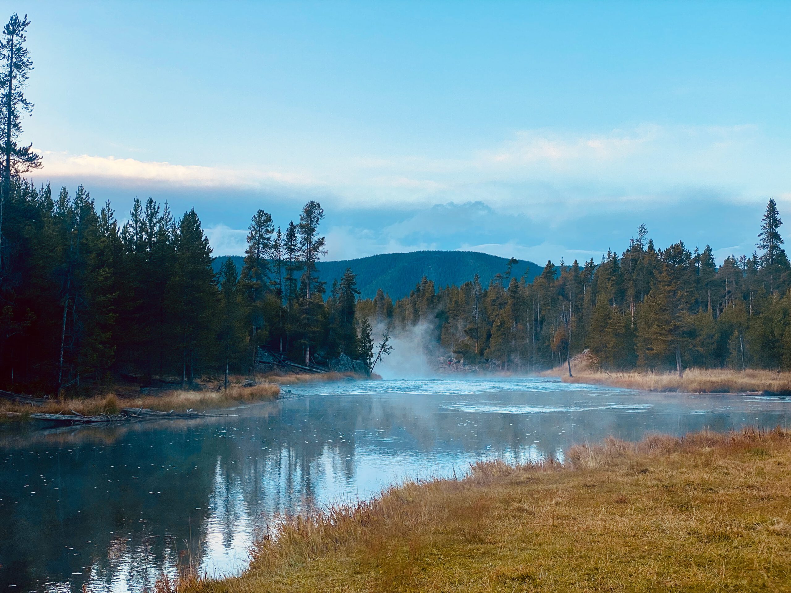 Calm geothermal river at sunrise in Yellowstone National Park, with steam rising above the water, pine forests along the riverbanks, and mountains in the background reflecting on the surface.