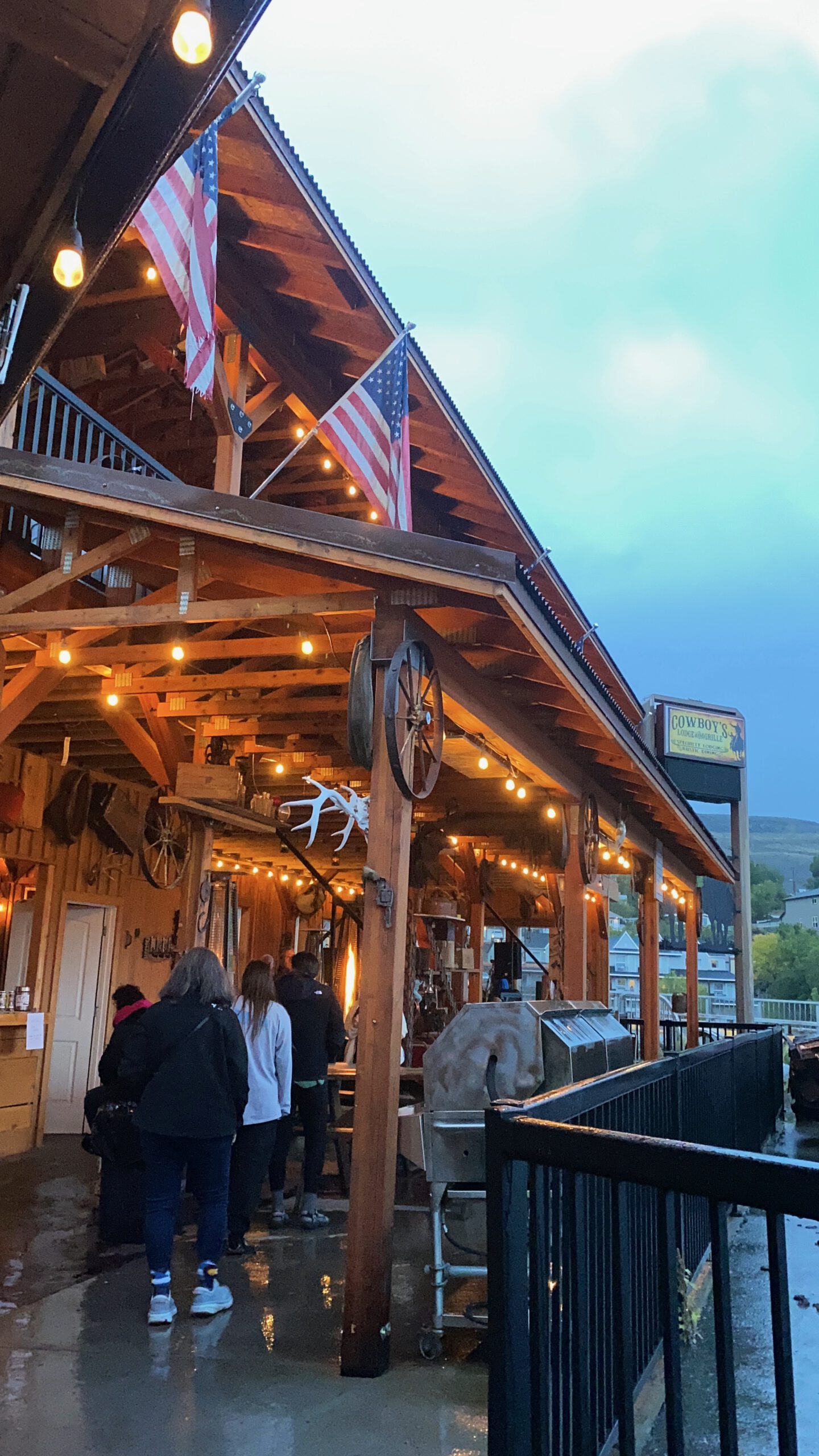Outdoor patio at Cowboy’s Lodge and Grill in Gardiner, Montana with American flags and warm string lights and live country music event in front of the lodge