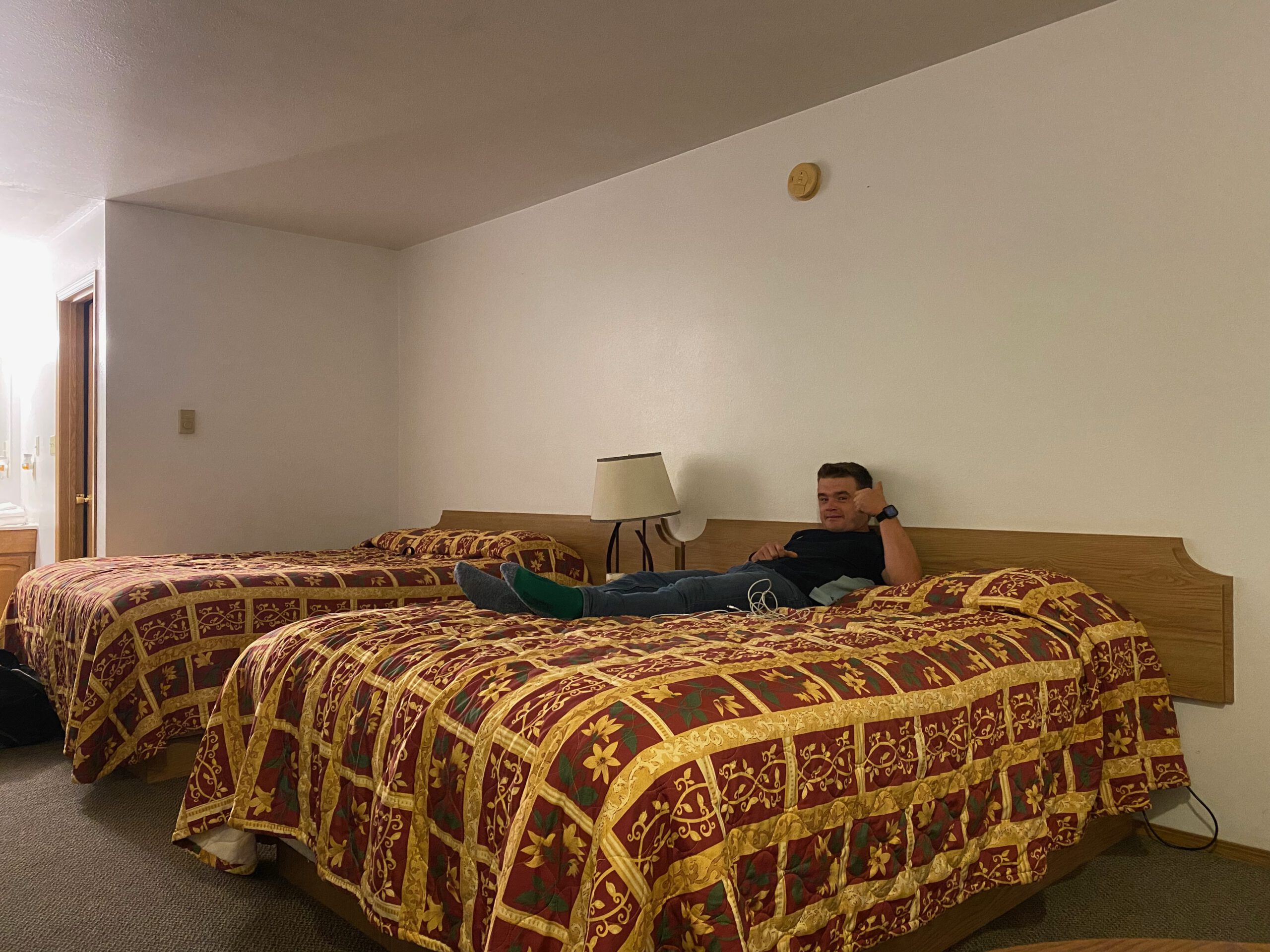 Interior of a double room at Arco Inn Motel in Idaho, featuring two queen beds with red and gold patterned bedspreads decorated with floral and geometric motifs, a simple wooden headboard, bedside lamp, and classic American motel atmosphere