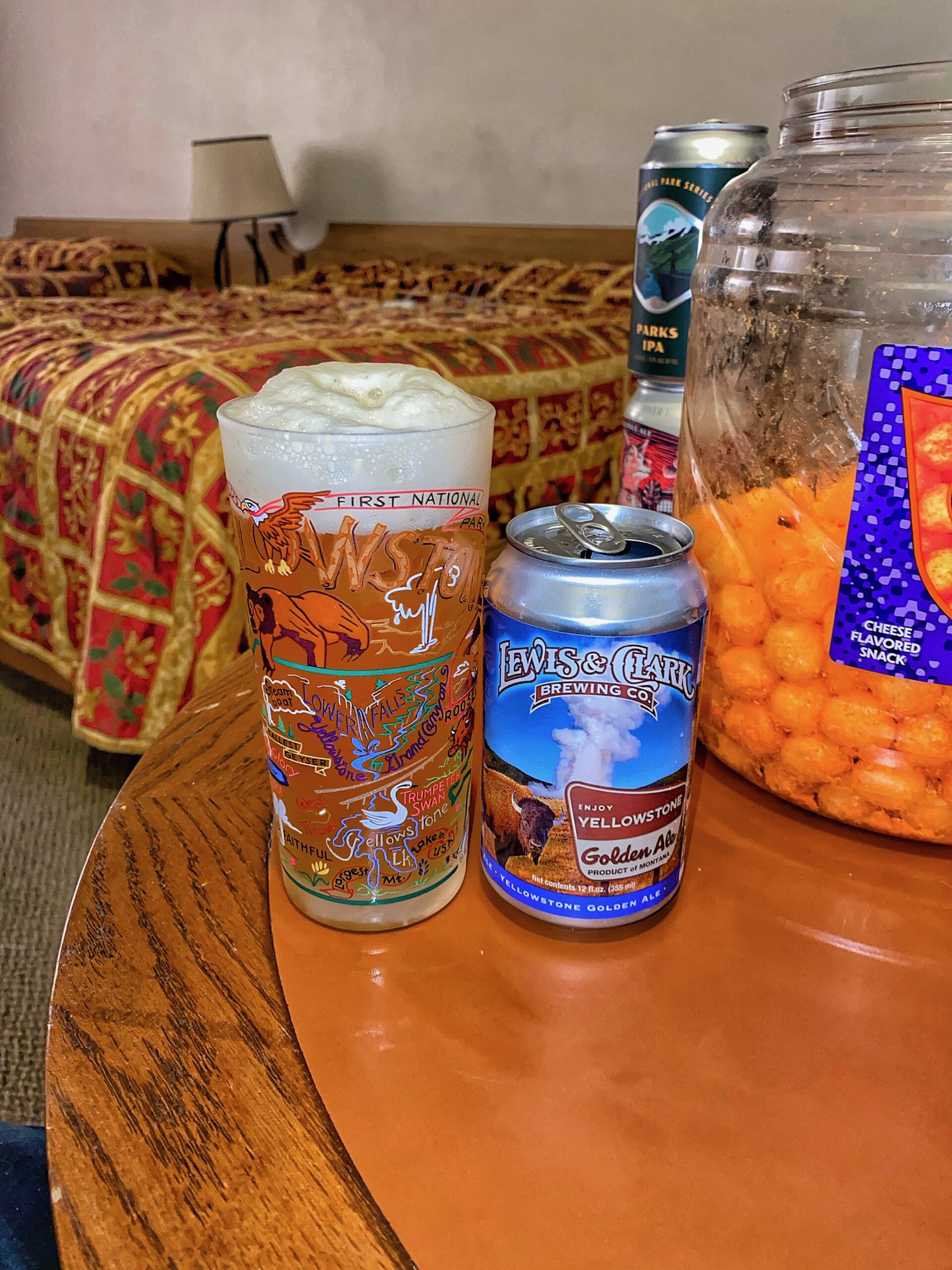 Lewis and Clark Brewing Co Yellowstone Golden Ale served in a Yellowstone National Park themed beer glass next to a jar of cheese flavored snacks inside an Arco Inn Motel room, with classic red and gold patterned motel bedspread in the background