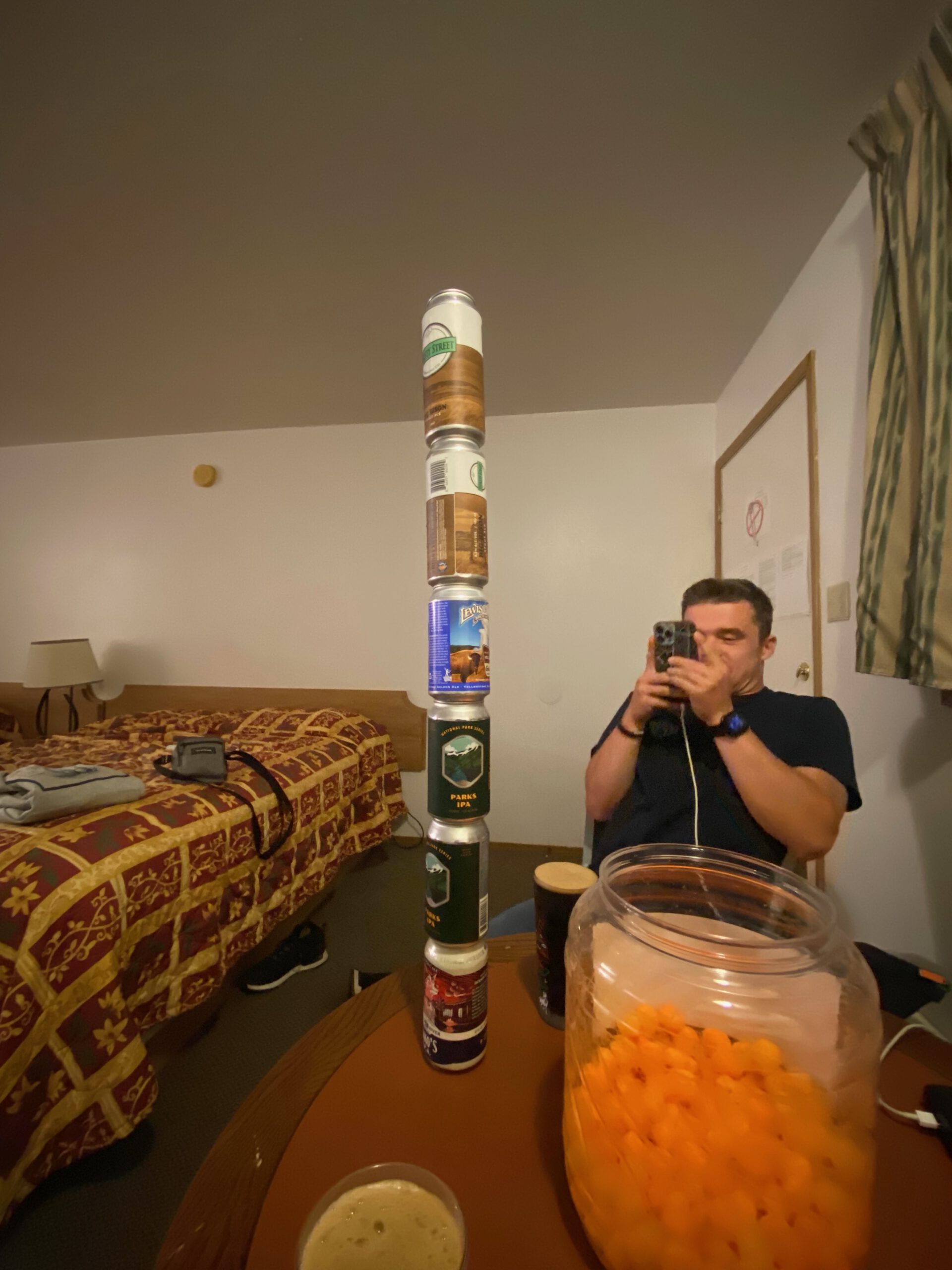 Stack of empty craft beer cans including Lewis and Clark Brewing Co and national park themed beers balanced on a table inside an American motel room, capturing a relaxed road trip night atmosphere