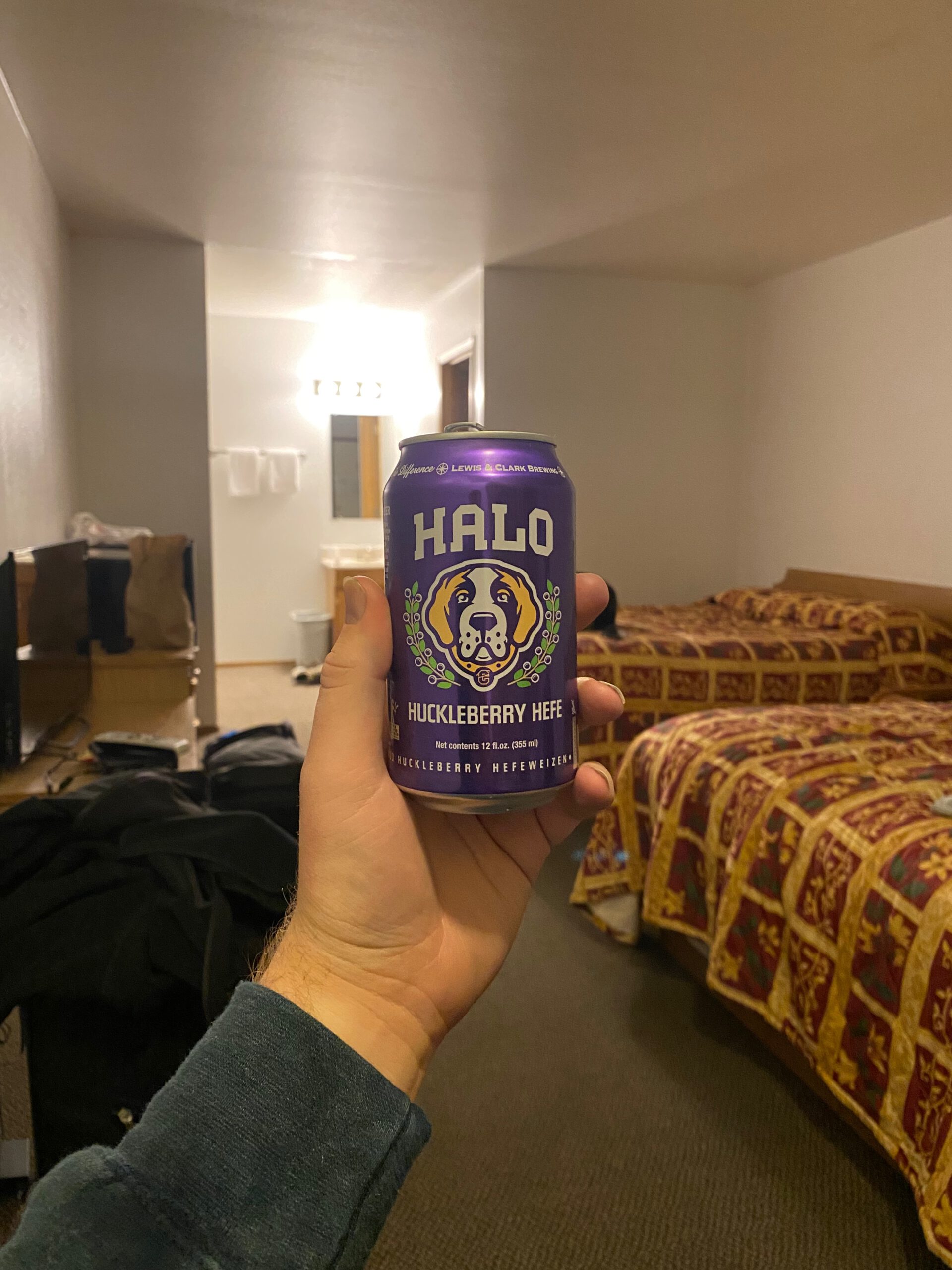 Halo Huckleberry Hefe beer can from Lewis and Clark Brewing Co held inside an American motel room at Arco Inn Motel, with classic patterned motel beds in the background during a road trip evening