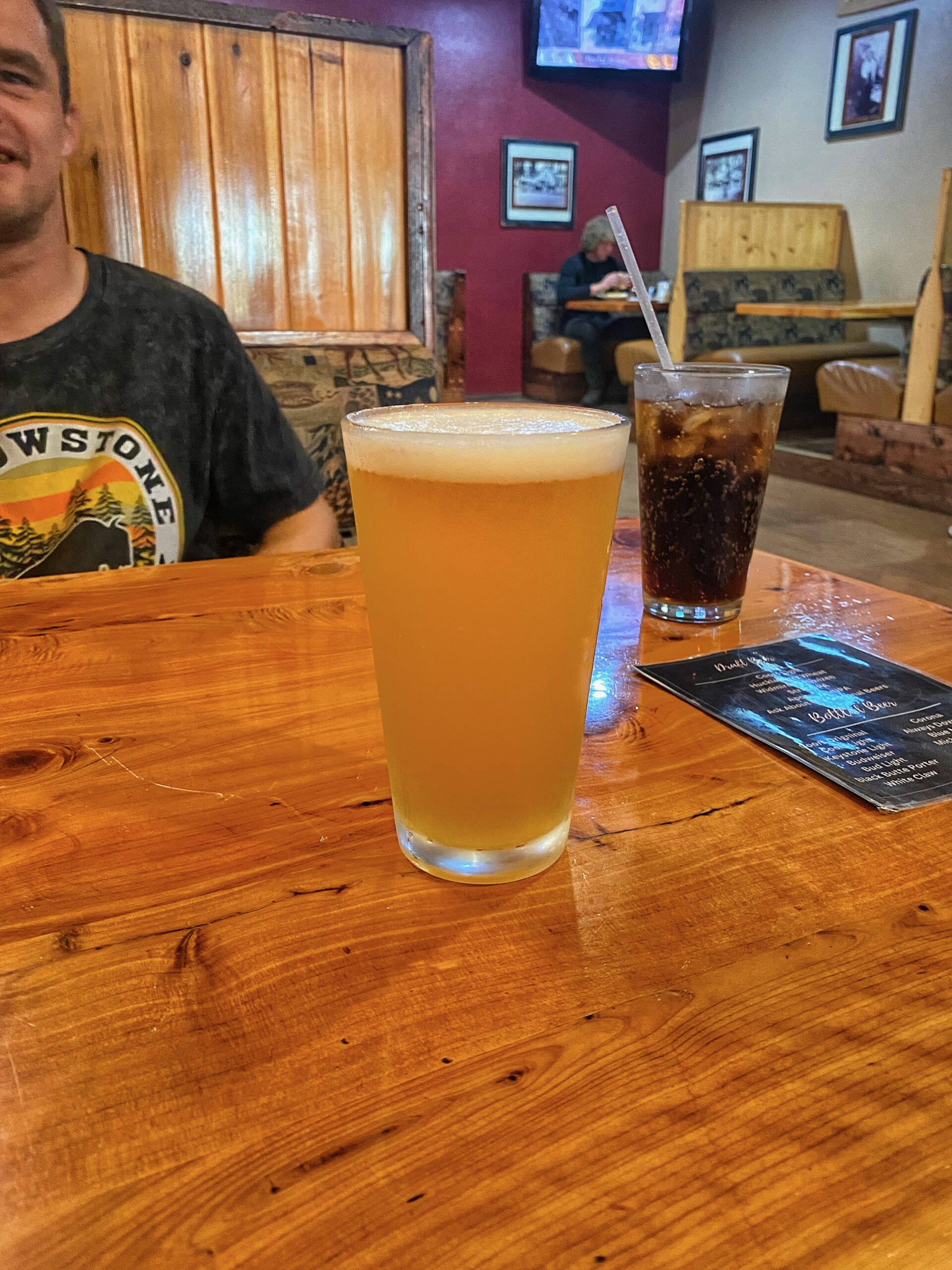 Cold draft beer and a glass of soda on a wooden table inside Outpost Pizza Pub & Grill John Day, with rustic booth seating, warm interior lighting, and a casual American pub atmosphere, showing the relaxed dining experience in John Day, Oregon.