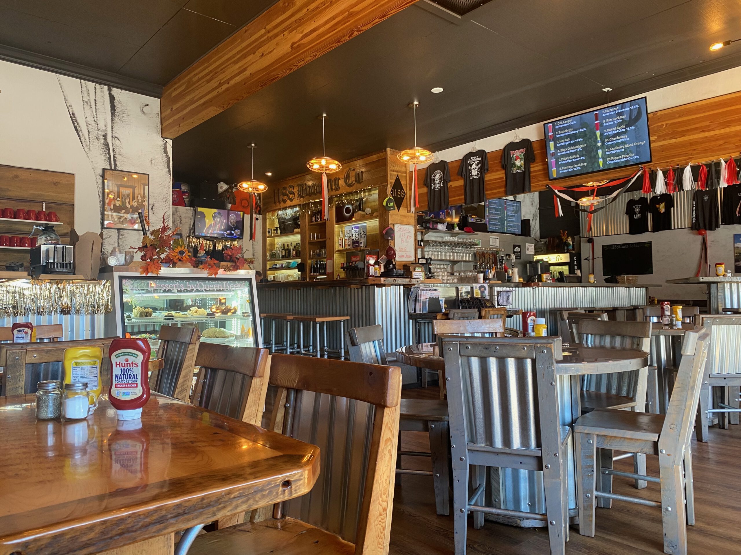 Spacious interior of 1188 Brewing Company in John Day Oregon, showcasing rustic wooden tables, metal accents, warm hanging lights, the main bar, and tap list screens in a welcoming small town craft brewery setting.