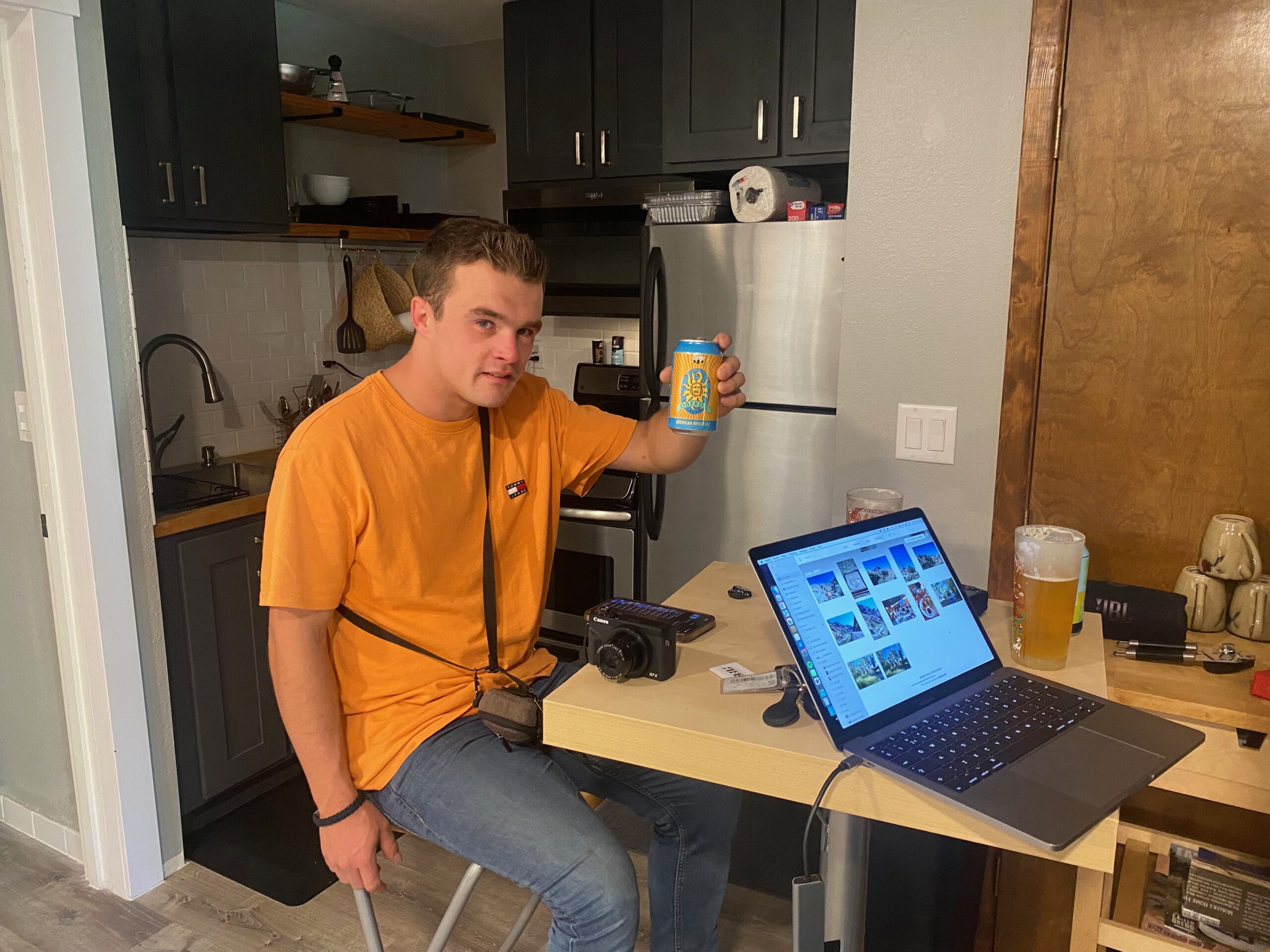 Peter Smit - peter010boy - sitting at the kitchen table inside the Airbnb John Day Oregon, holding a can of beer, with a MacBook Pro 2017 open showing travel photos, a compact camera on the table, and a small modern kitchen visible in the background during a relaxed evening indoors.