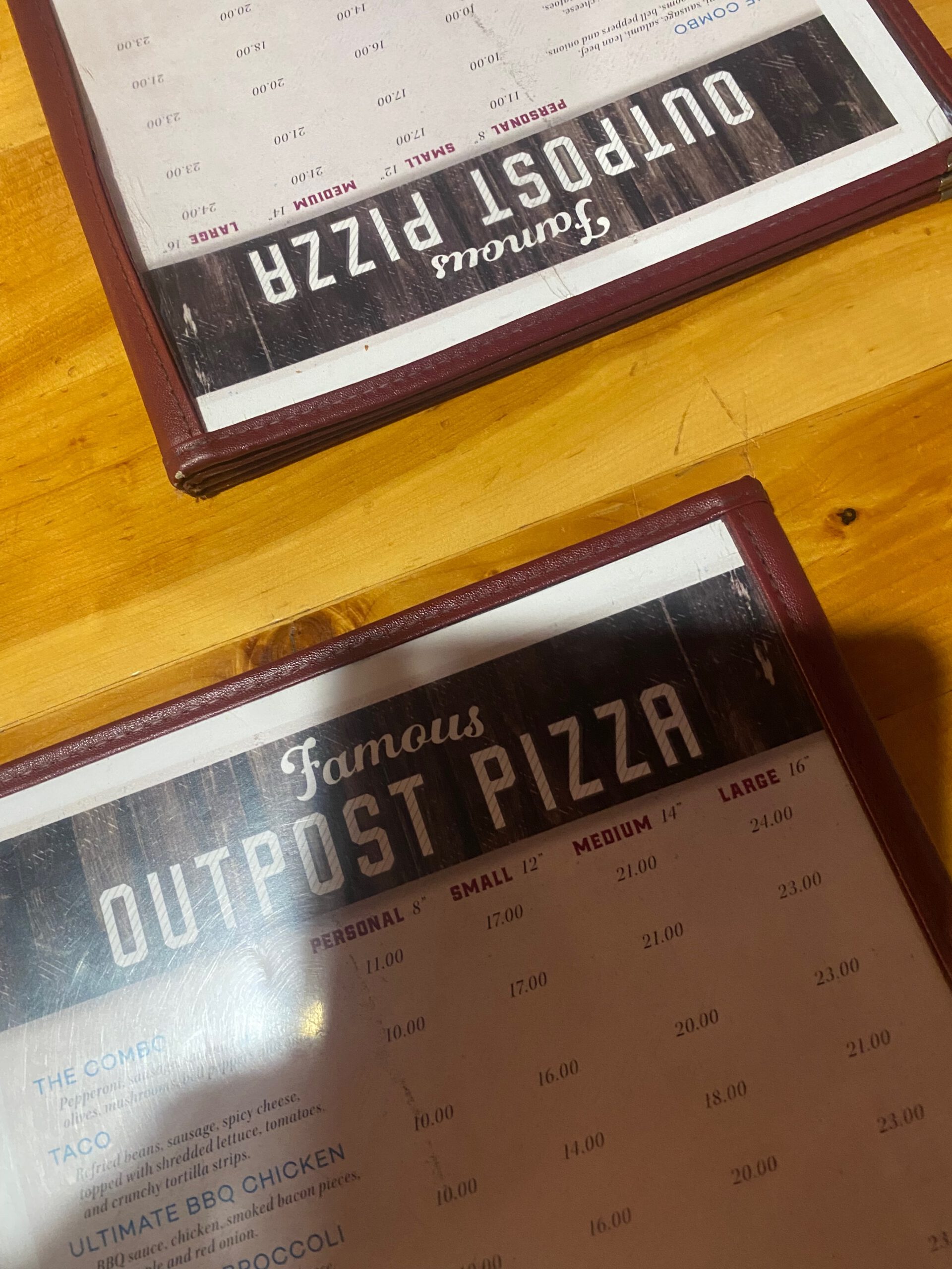 Close-up of menu cards at Outpost Pizza Pub & Grill John Day, showing the famous Outpost pizza menu with sizes and prices, placed on a wooden table inside the restaurant in John Day, Oregon.