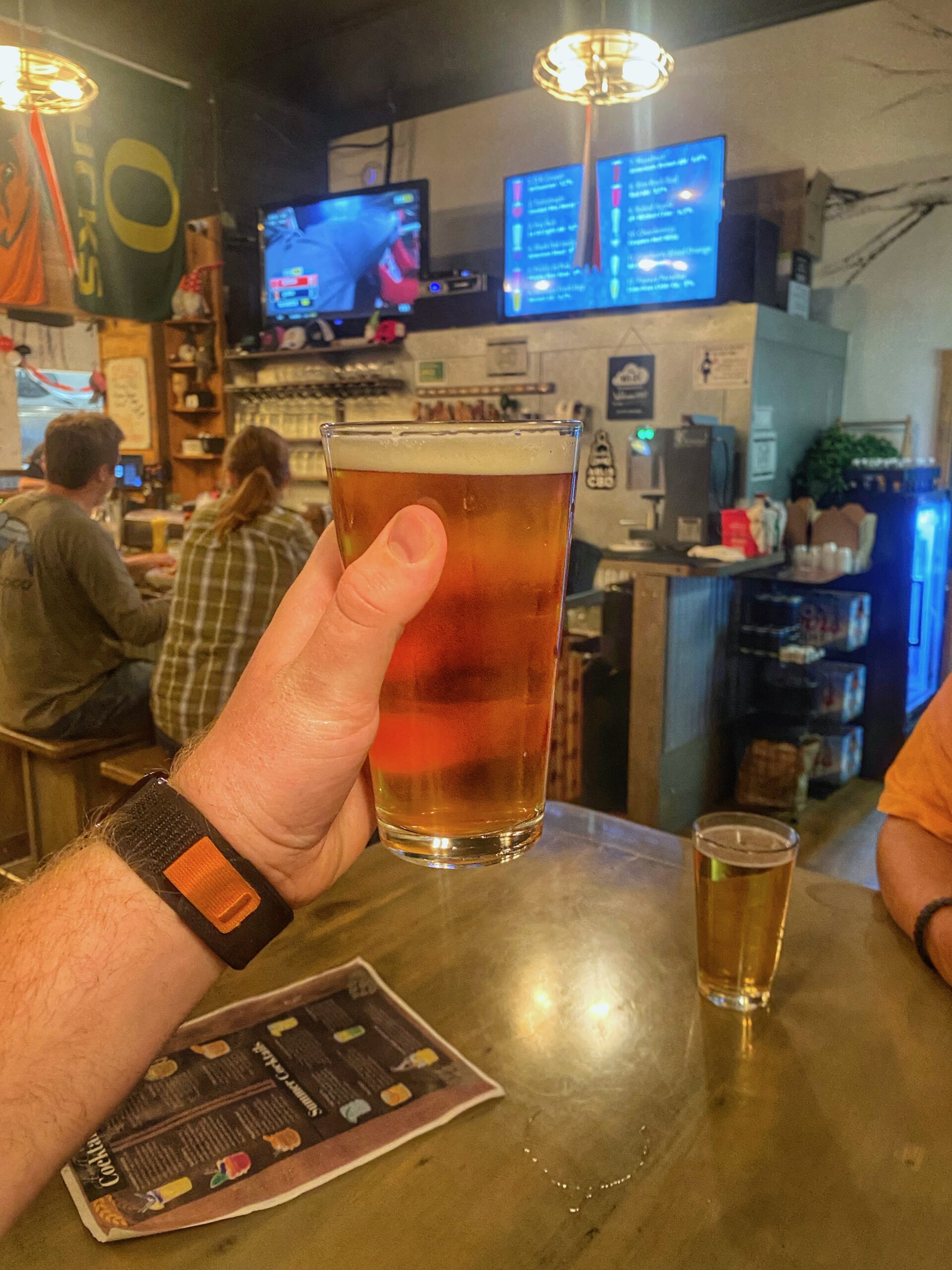 Hand holding a pint of golden craft beer inside 1188 Brewing Company in John Day Oregon, with the bar area and tap list screens visible in the background of a cozy small town brewery.