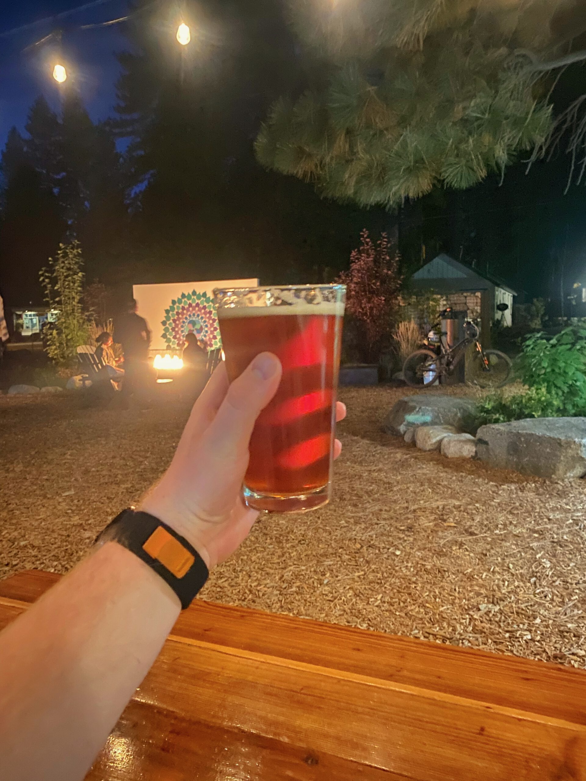 Hand holding a Tahoe Red Ale 5.4 percent at Bear Belly Brewing Company in Kings Beach California, with outdoor seating, fire pit, and evening atmosphere at a Lake Tahoe brewery