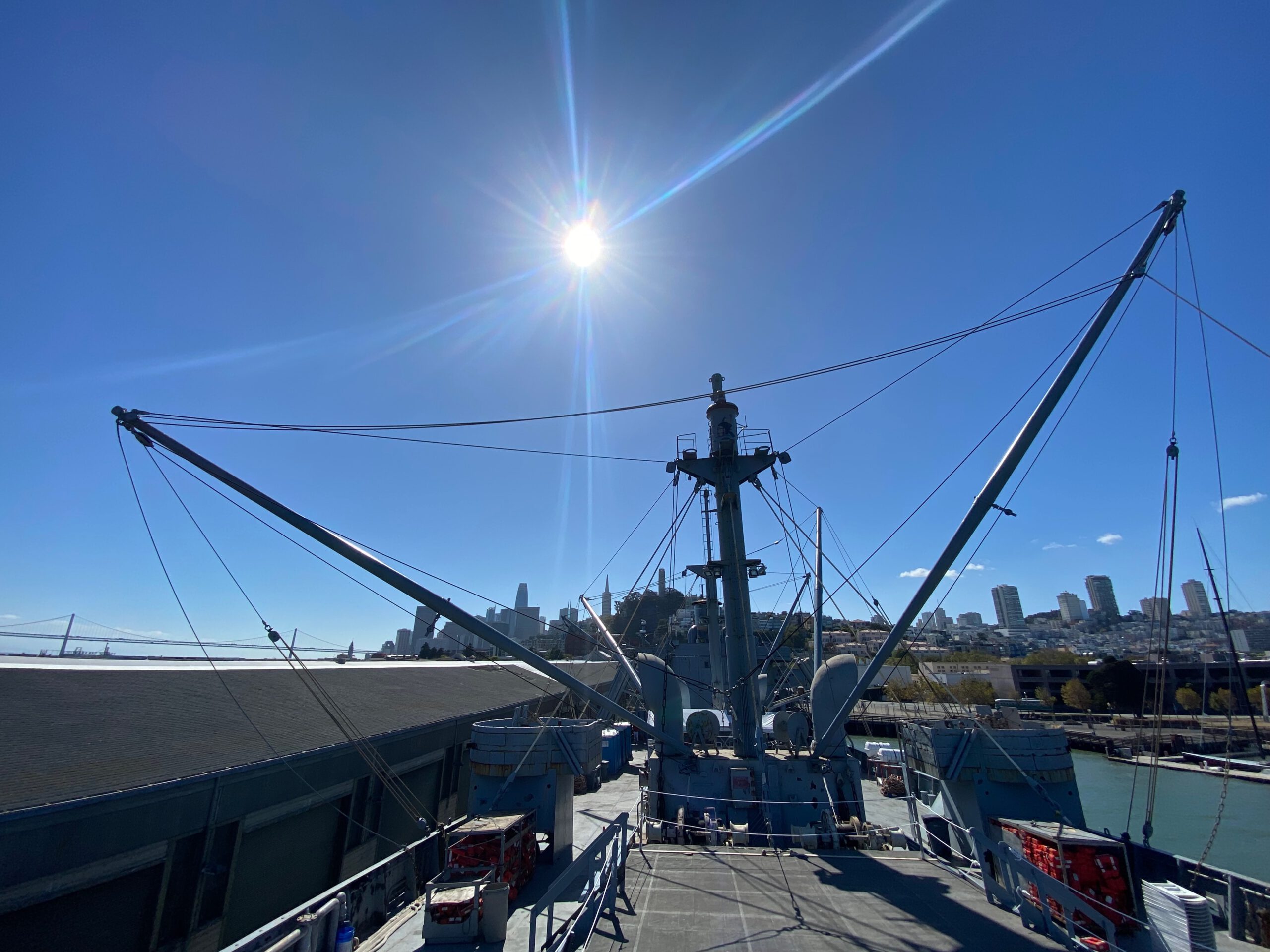 View across the main deck of the SS Jeremiah O'Brien San Francisco under a bright California sun, with cargo cranes, rigging, and the San Francisco skyline visible in the background.